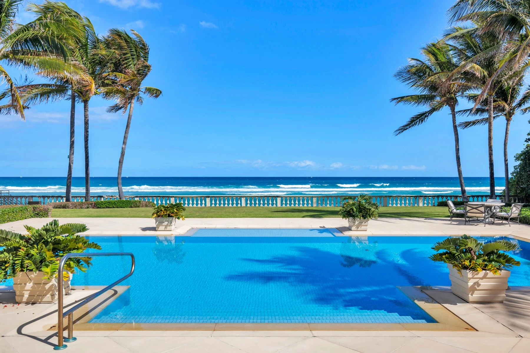 Swimming pool with ocean view, palm trees, and outdoor seating on a bright sunny day.
