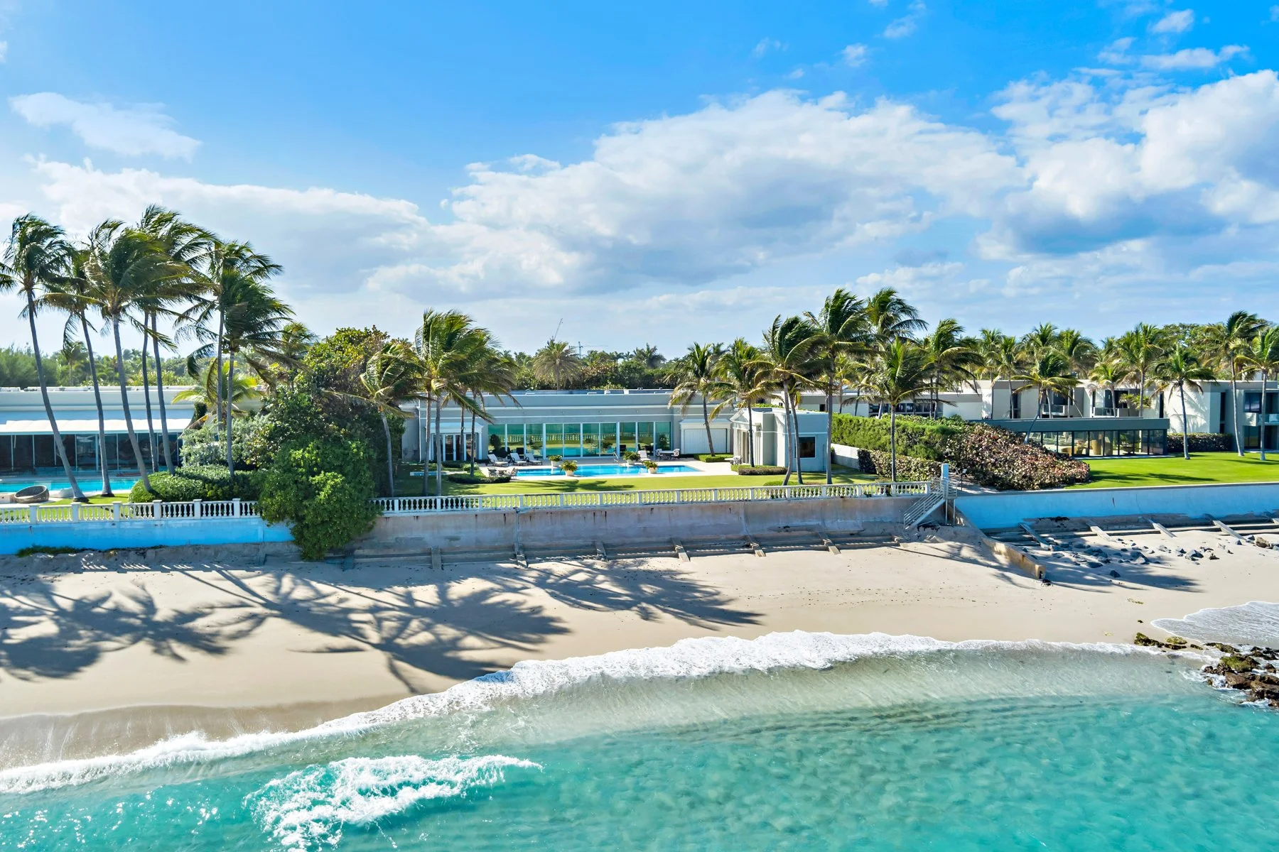 Luxury beachfront property with swimming pool, palm trees, and ocean in the foreground under a partly cloudy sky.