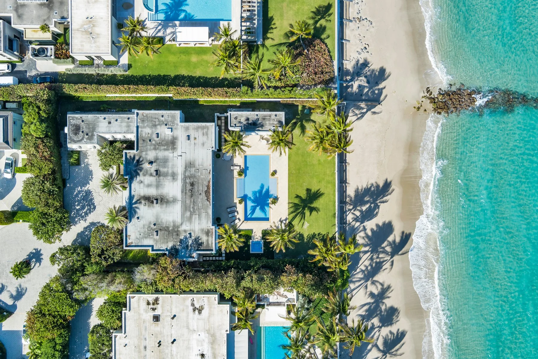 An aerial view of a beachfront property with a swimming pool, palm trees, green lawn, and white sand beach with turquoise water.