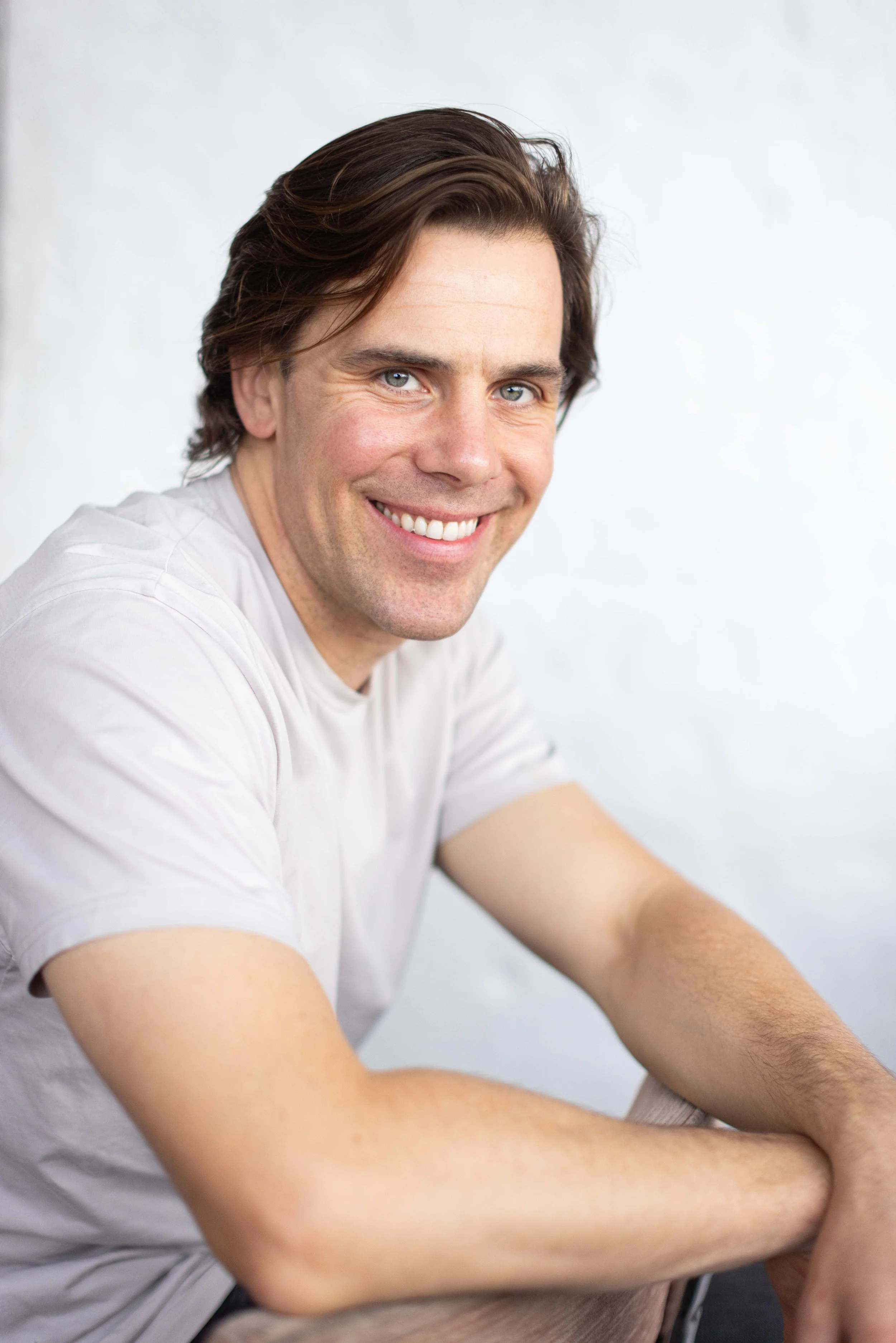 Close-up of a smiling man with brown hair and blue eyes, wearing a white t-shirt, sitting with his arms resting on a surface against a plain light gray background.