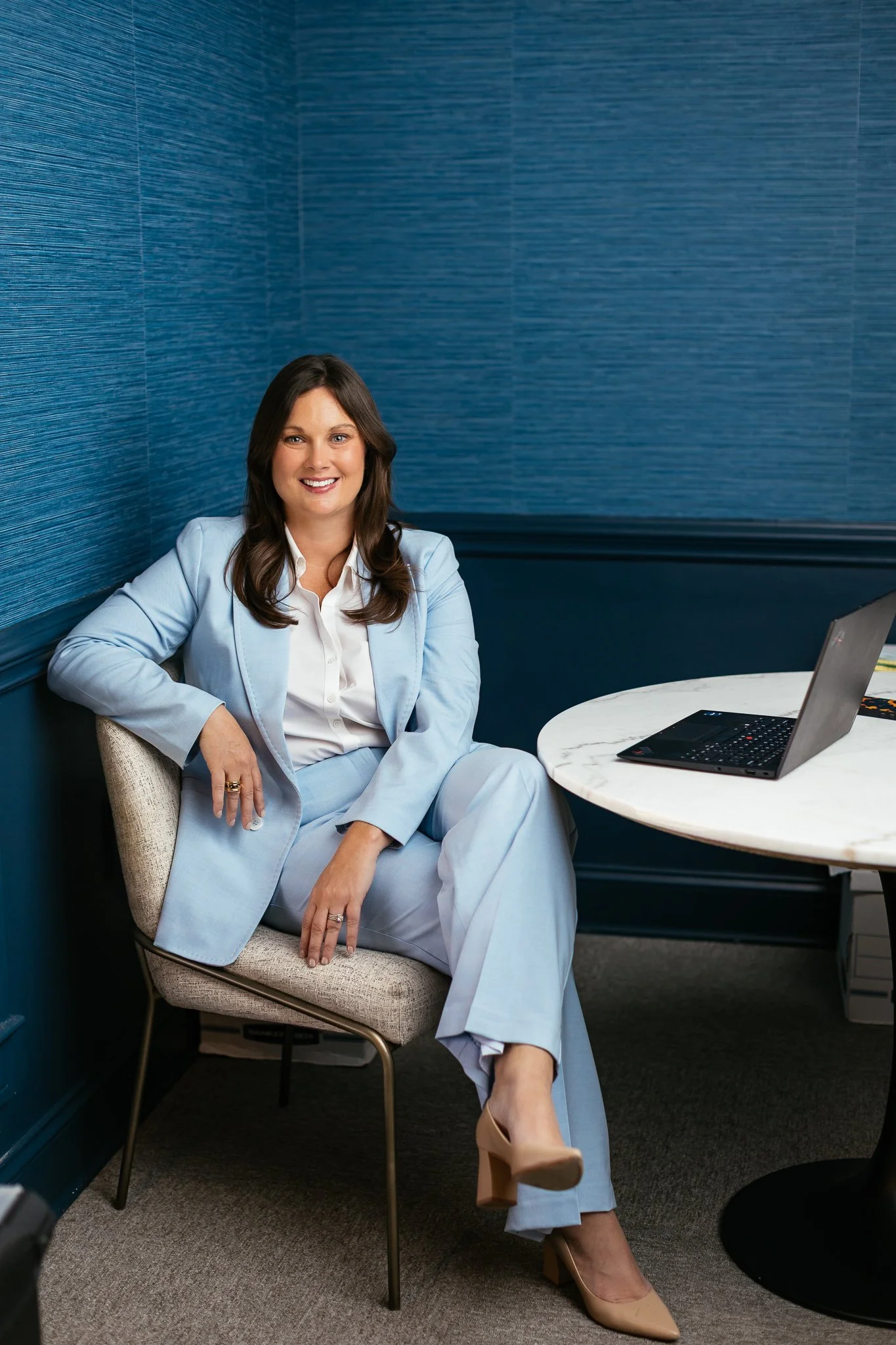 A woman in a light blue business suit sitting in a chair next to a round table with a laptop on it, smiling at the camera.