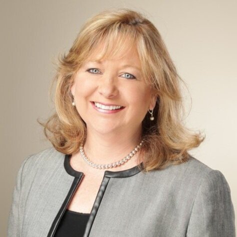 Professional woman with blonde hair smiling, wearing a gray blazer, pearl necklace, and pearl earrings, against a neutral background.