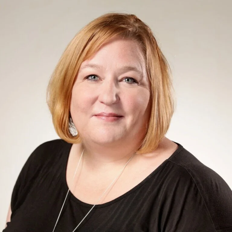 Portrait of a woman with shoulder-length red hair, wearing a black top and a necklace, smiling softly at the camera.