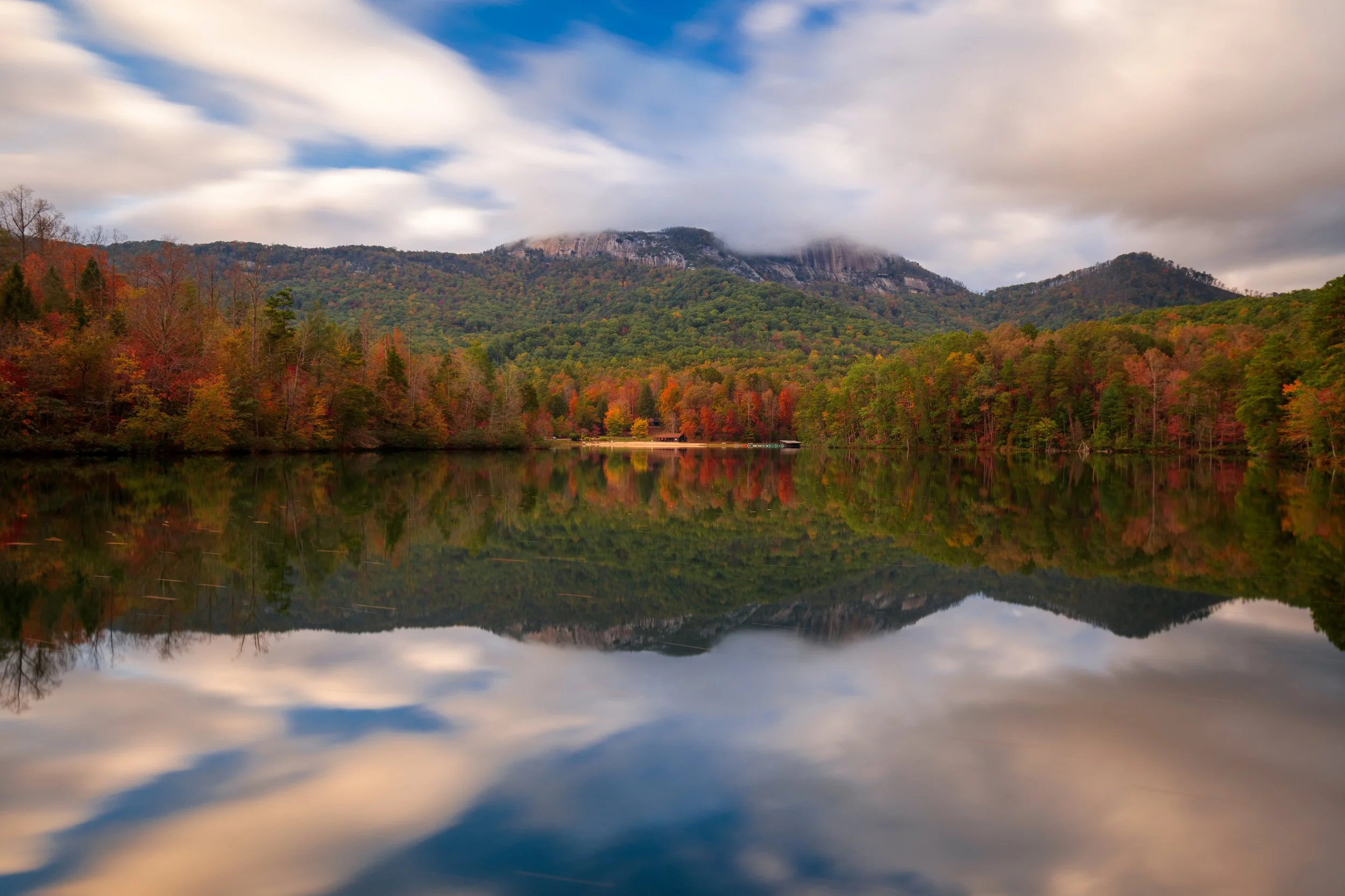 Scenic view of a calm lake reflecting a forest with autumn-colored trees and mountains in the background under a partly cloudy sky.
