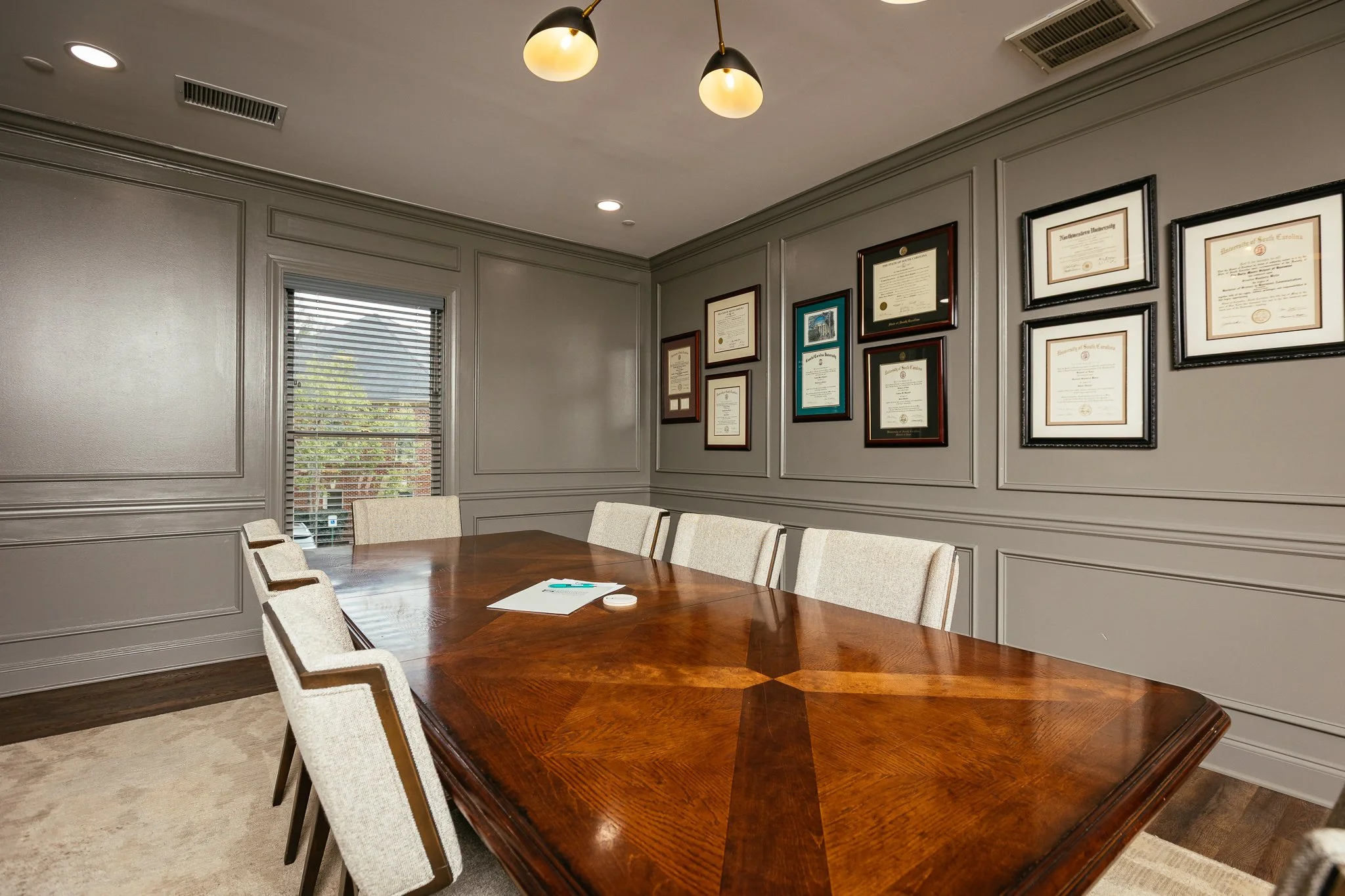 A meeting room with a large polished wooden table surrounded by beige upholstered chairs. The walls are gray with framed certificates and diplomas. A window with blinds is visible, with sunlight coming through.