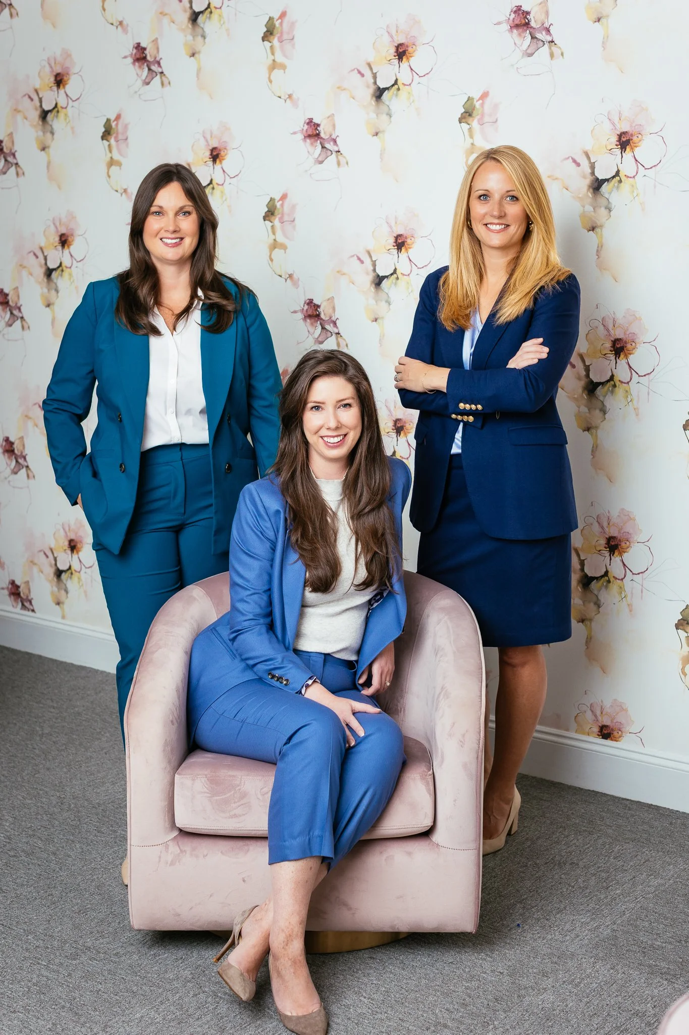 Three women in professional attire posing indoors against a floral wallpaper background. One woman is seated on a pink armchair, and two women are standing on either side, smiling at the camera.