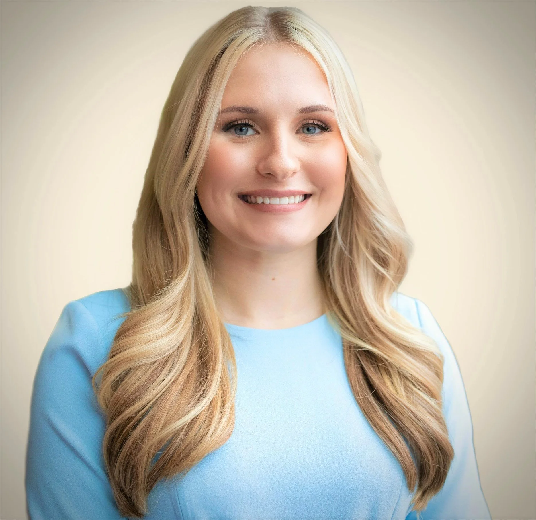 A portrait of a young woman with long blonde hair wearing a light blue top, smiling against a neutral background.