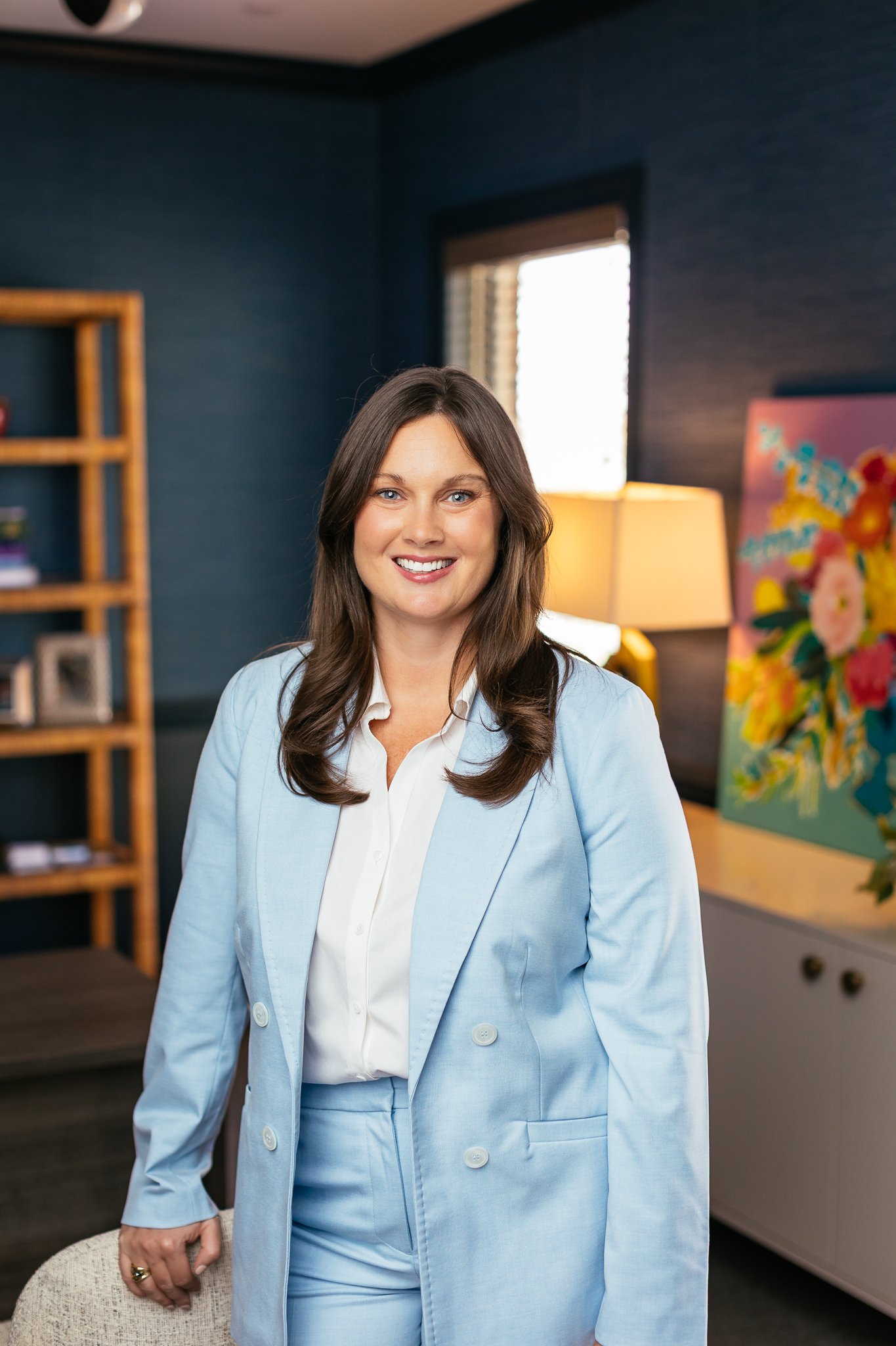 A woman with long brown hair smiling, wearing a light blue suit in an office with dark walls, a bookshelf, a lamp, and a colorful painting.
