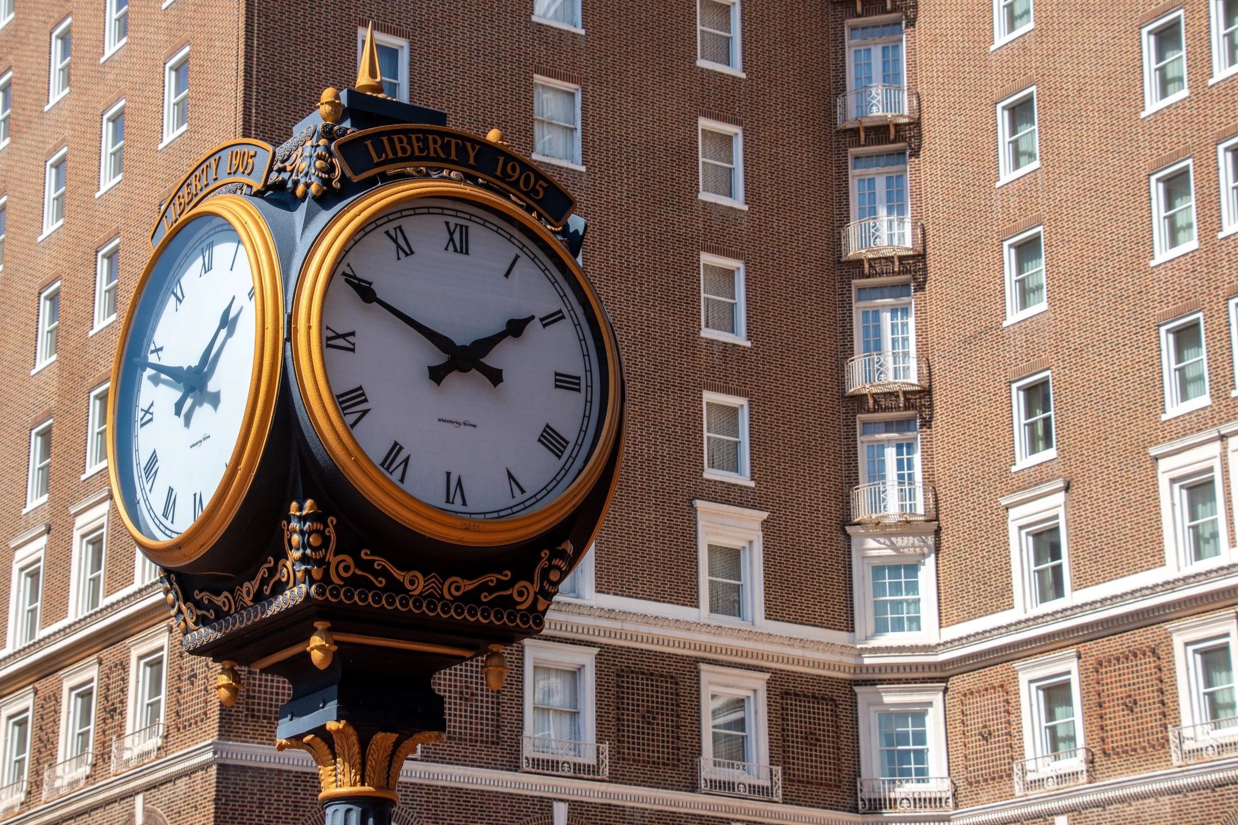 A vintage clock with black and gold accents, showing the time as 2:53, located outside against a background of a brick building with multiple windows and small balconies.