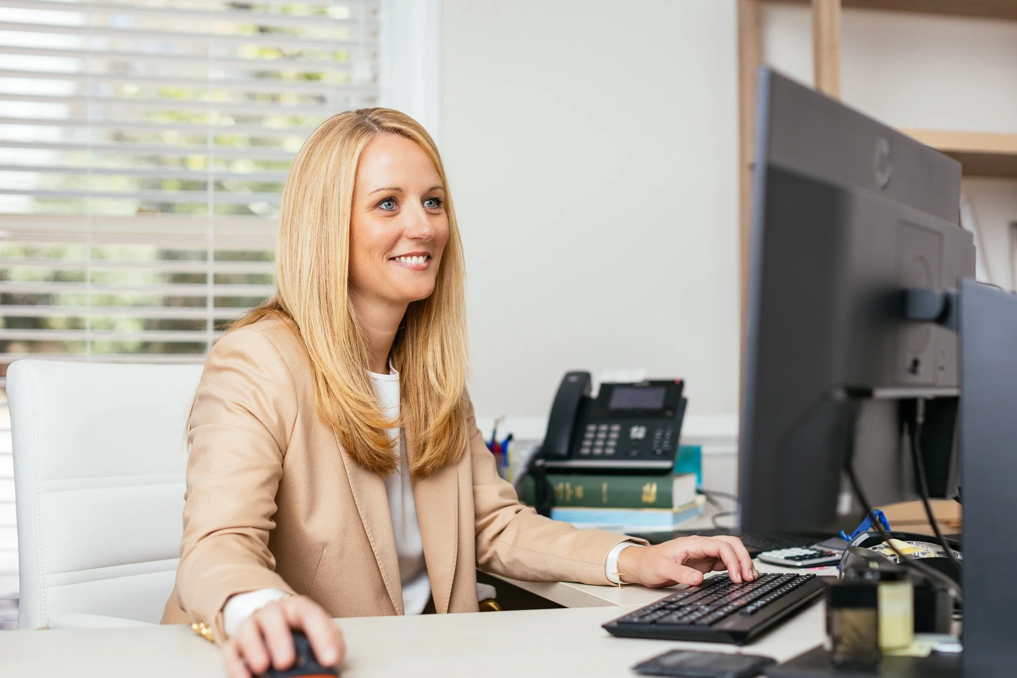 A woman with blonde hair, dressed in a beige blazer, working at her desk with a computer, phone, and office supplies in an office setting.
