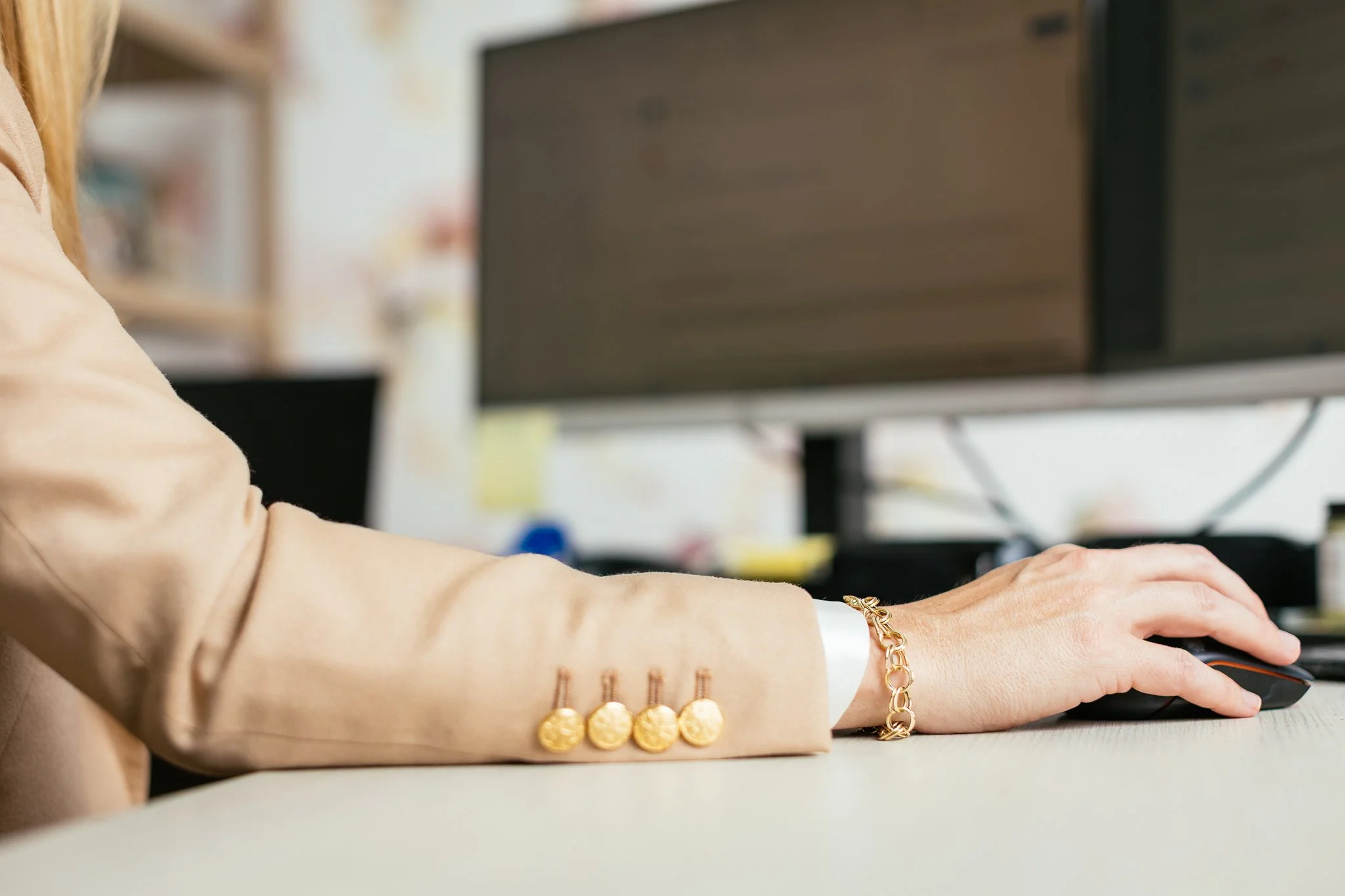Close-up of a person's arm using a computer mouse on a desk, wearing a beige blazer with gold jewelry, in an office setting with computer monitors in the background.
