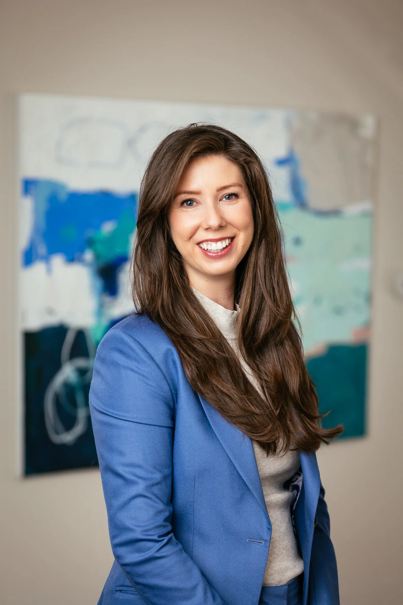 A smiling woman with long brown hair wearing a blue blazer and gray top, standing in front of an abstract painting with blue, green, and white colors.