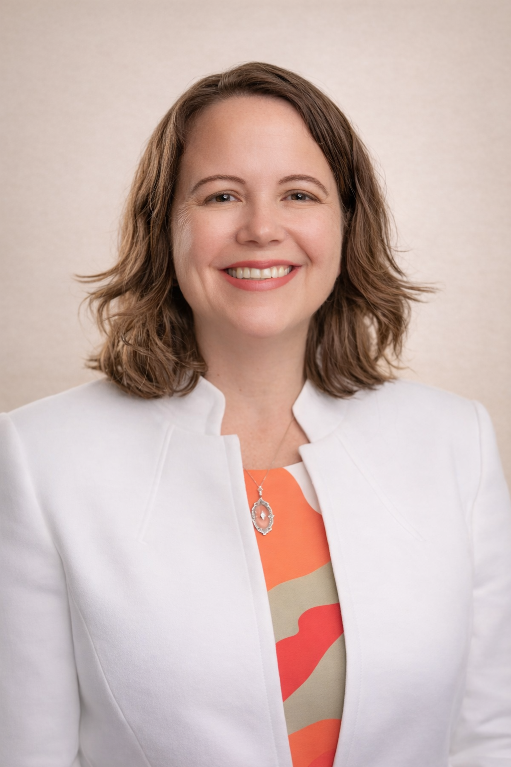 A smiling woman with shoulder-length brown hair, wearing a white blazer and a colorful top, standing in what appears to be an office setting.