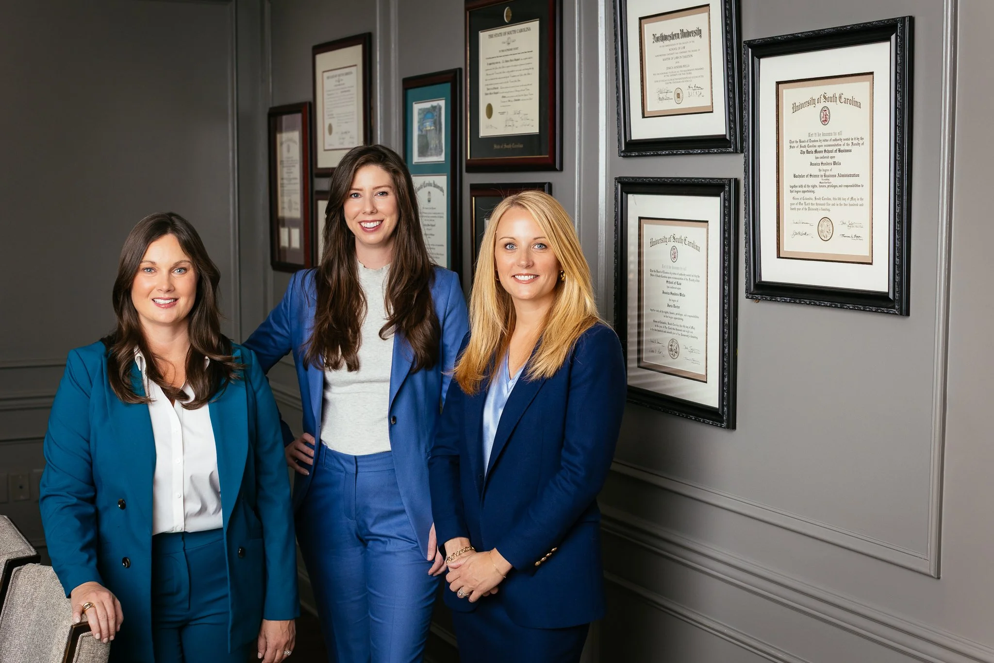 Three women dressed in blue suits standing in front of a wall with framed certificates and degrees, smiling at the camera.