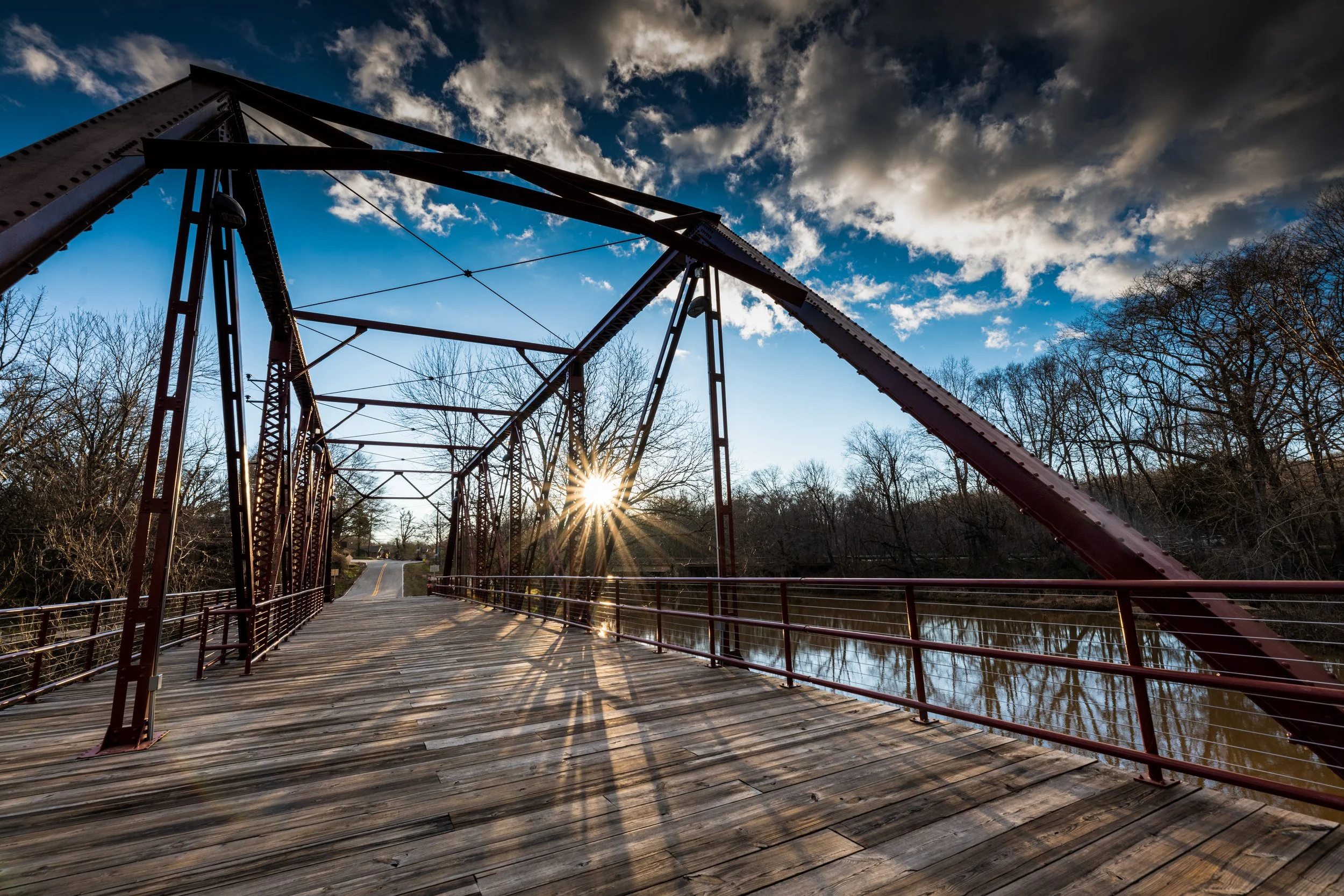 A wooden suspension bridge over a river with leafless trees on either side, the sun setting behind the bridge, creating a starburst effect in the sky with clouds.
