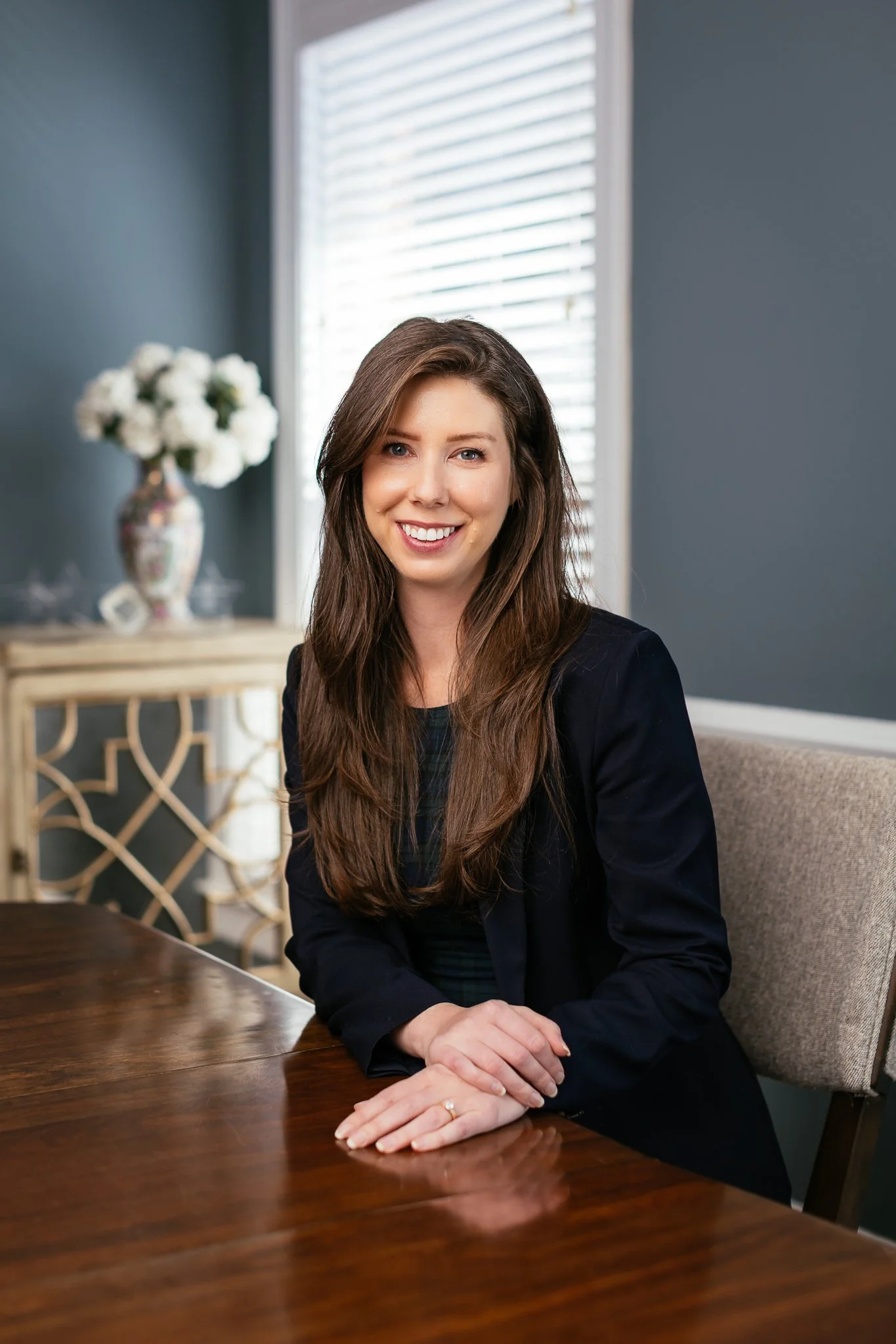 A woman with long brown hair and a black blazer, sitting at a wooden table, smiling at the camera with a vase of white flowers and a window with white blinds in the background.