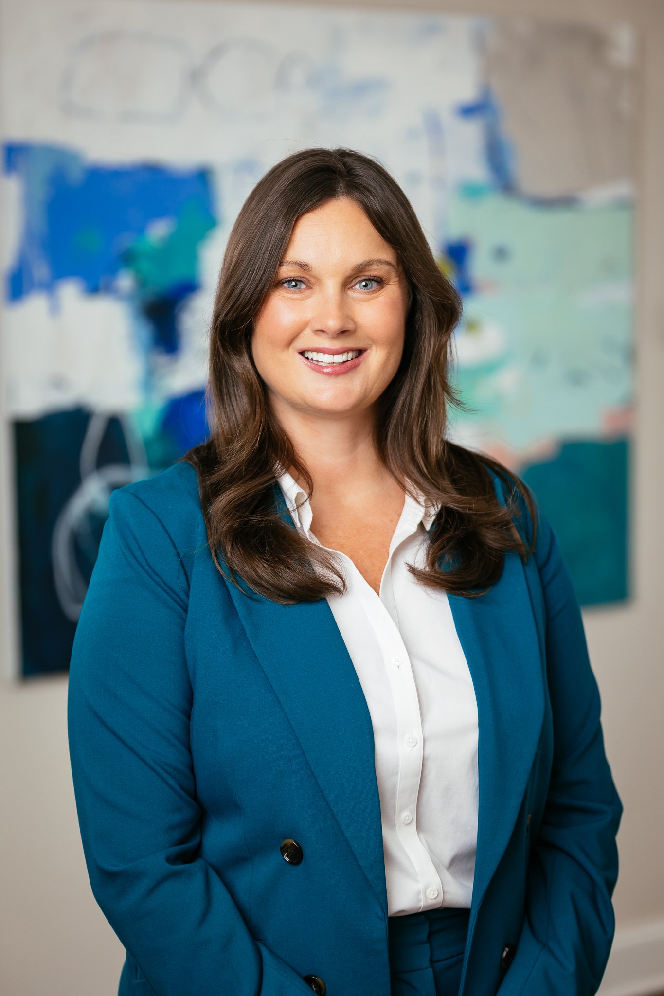 A woman with long brunette hair, wearing a blue blazer over a white shirt, smiling in front of an abstract painting.