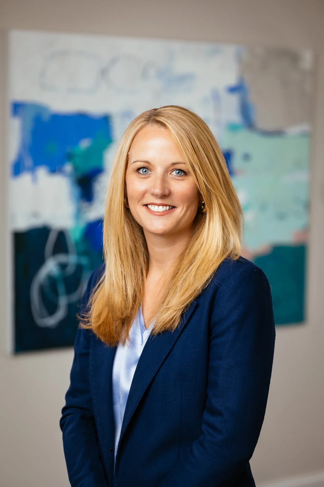 Professional woman with blonde hair smiling, wearing a navy blazer and light blue blouse, standing in front of an abstract painting.