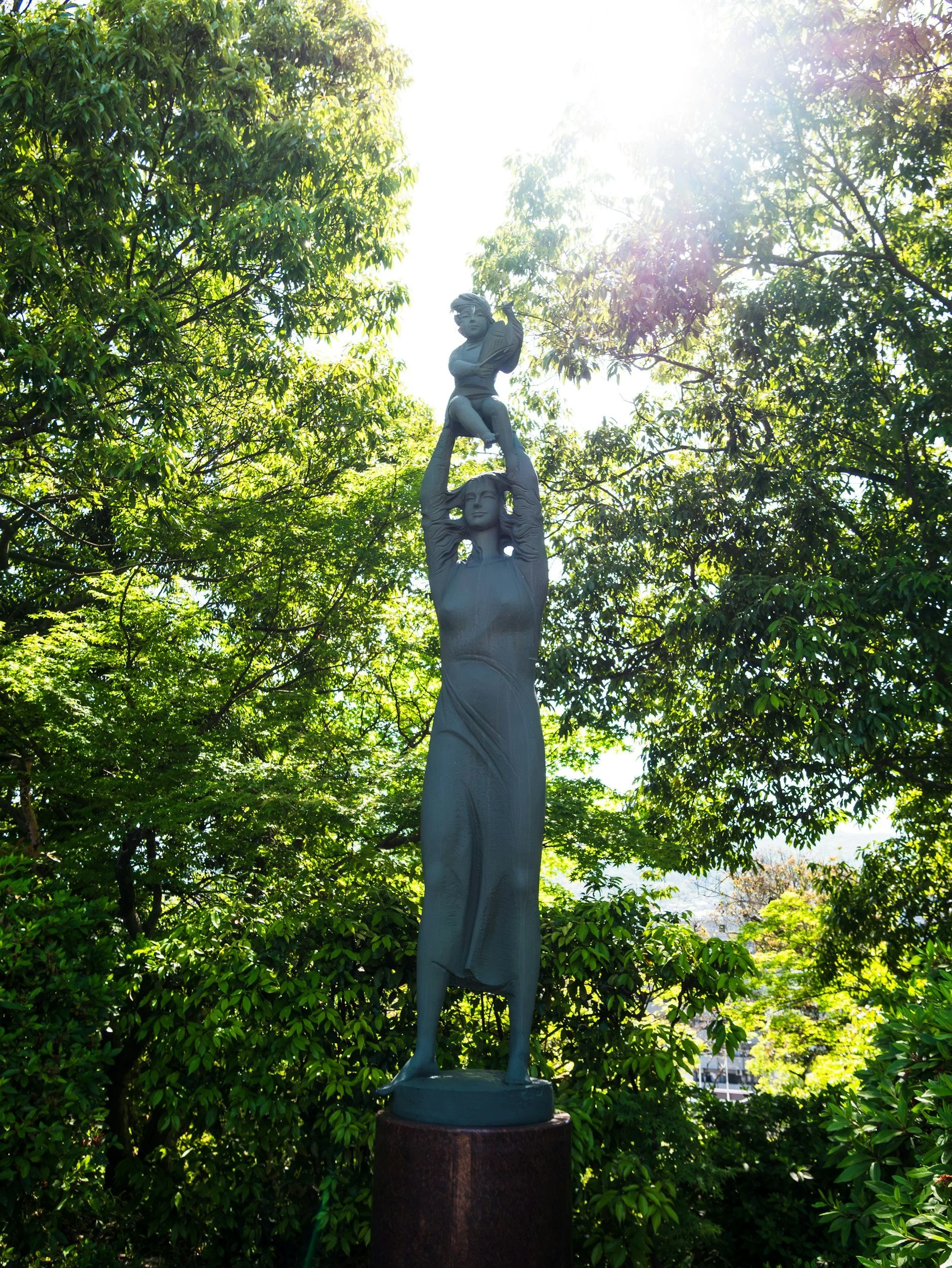 Skulptur av en kvinna som håller upp en pojke med en bok, placerad bland gröna träd med solljus som lyser starkt.