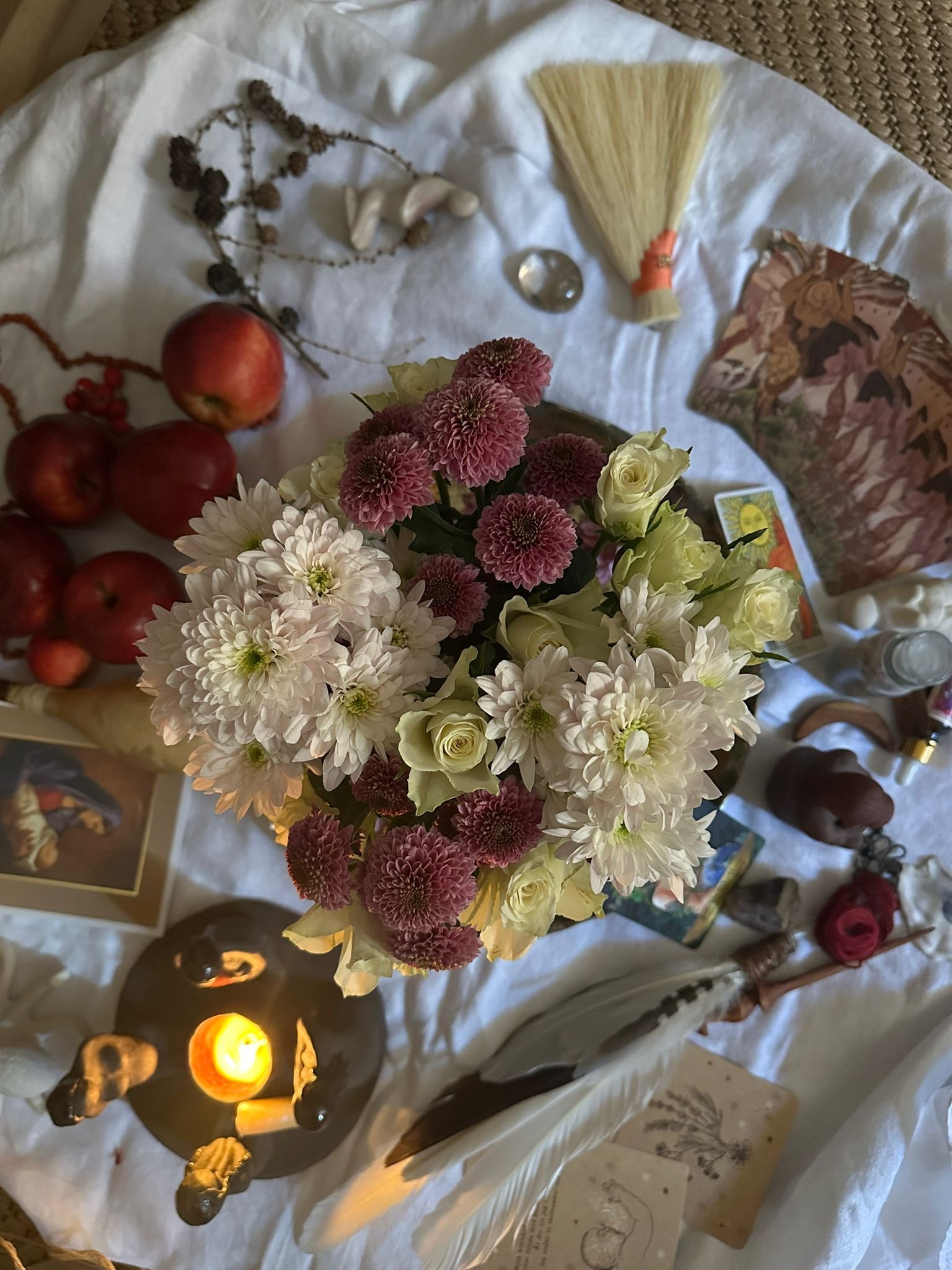 En bukett av blommor med rosa, vita och creamfärgade blommor, ett äpple, ett ljus i ett glas, ett litet silverföremål, och andra små föremål på ett vitt tyg.