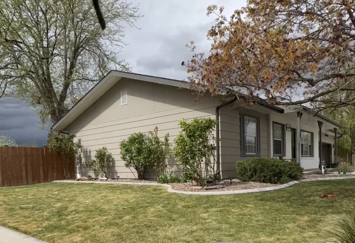 A single-story house with a beige exterior, gray trim, multiple windows, and a dark roof. There are bushes and trees in the front yard, with a brown wooden fence on the left. The sky is cloudy, with some dark storm clouds visible.