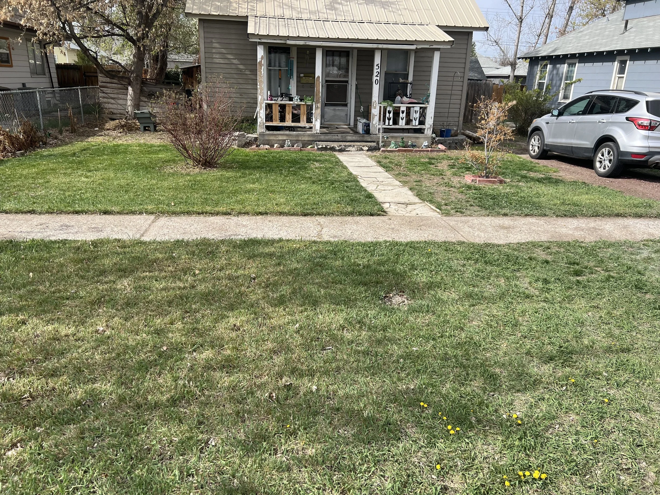 A small house with a covered porch, a house number 520, and a front yard with grass, a small pathway, and some bushes. A silver car is parked to the right side of the house.