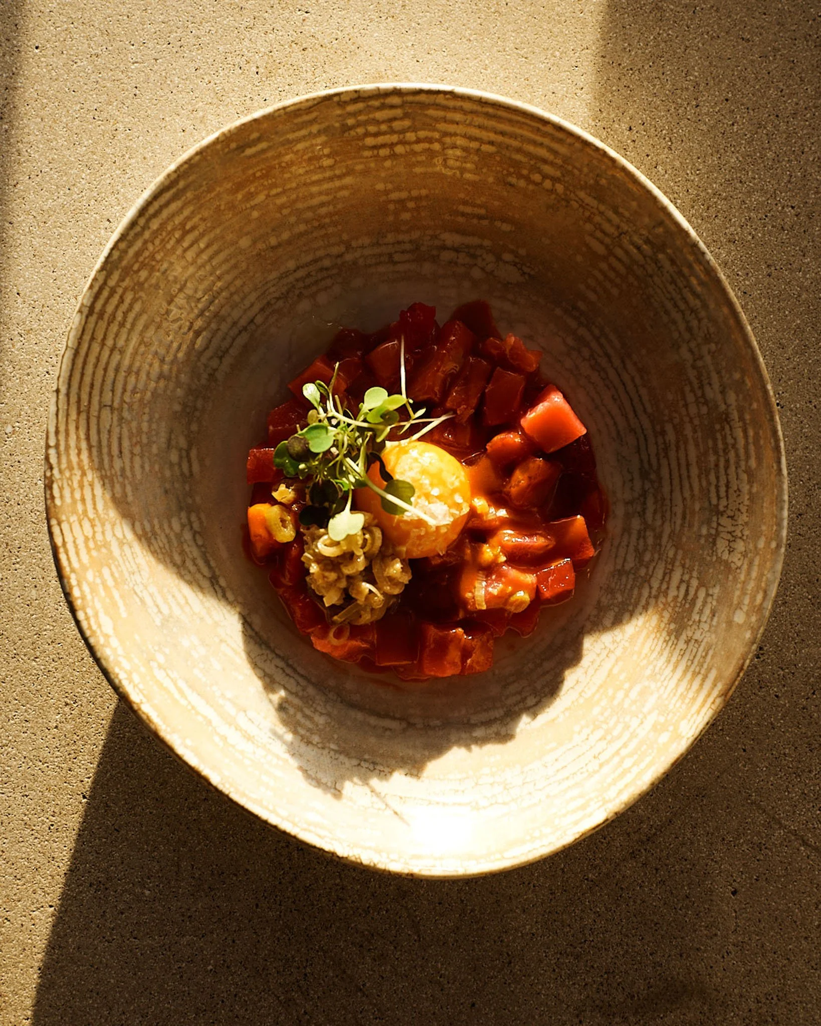 A bowl of raw beef cubes, garnished with microgreens, a dollop of yellow sauce, and a small pile of chopped vegetables, on a textured surface.