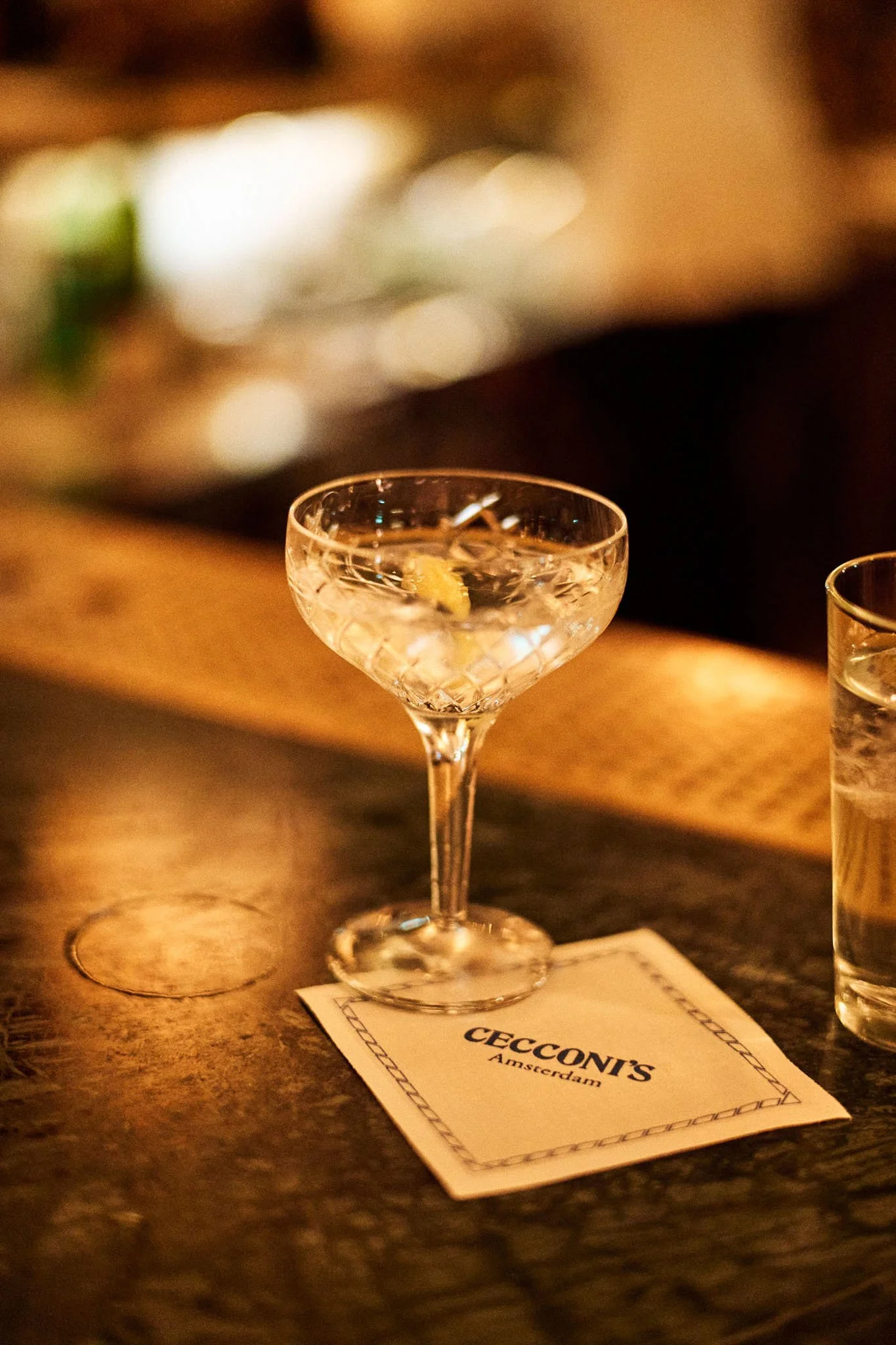 A cocktail glass on a wooden bar counter, filled with a clear drink and garnished with a lemon peel, next to a small glass of water, and a napkin with the logo 'Coconi's Amsterdam'.