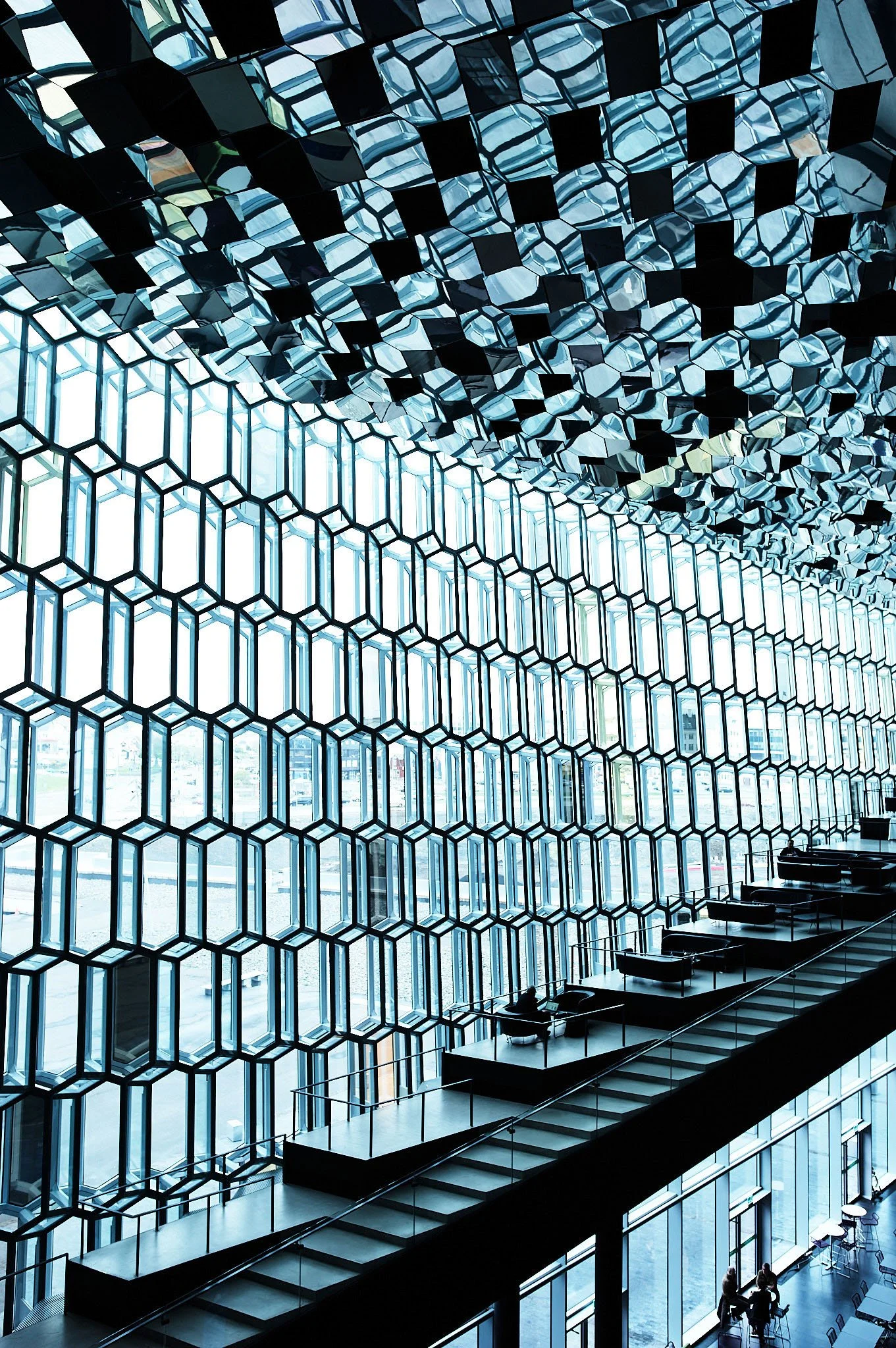 Interior of a modern building with a large hexagonal glass window wall and an artistic ceiling with mirrored hexagonal panels, featuring a mezzanine walkway with seating and a few people.