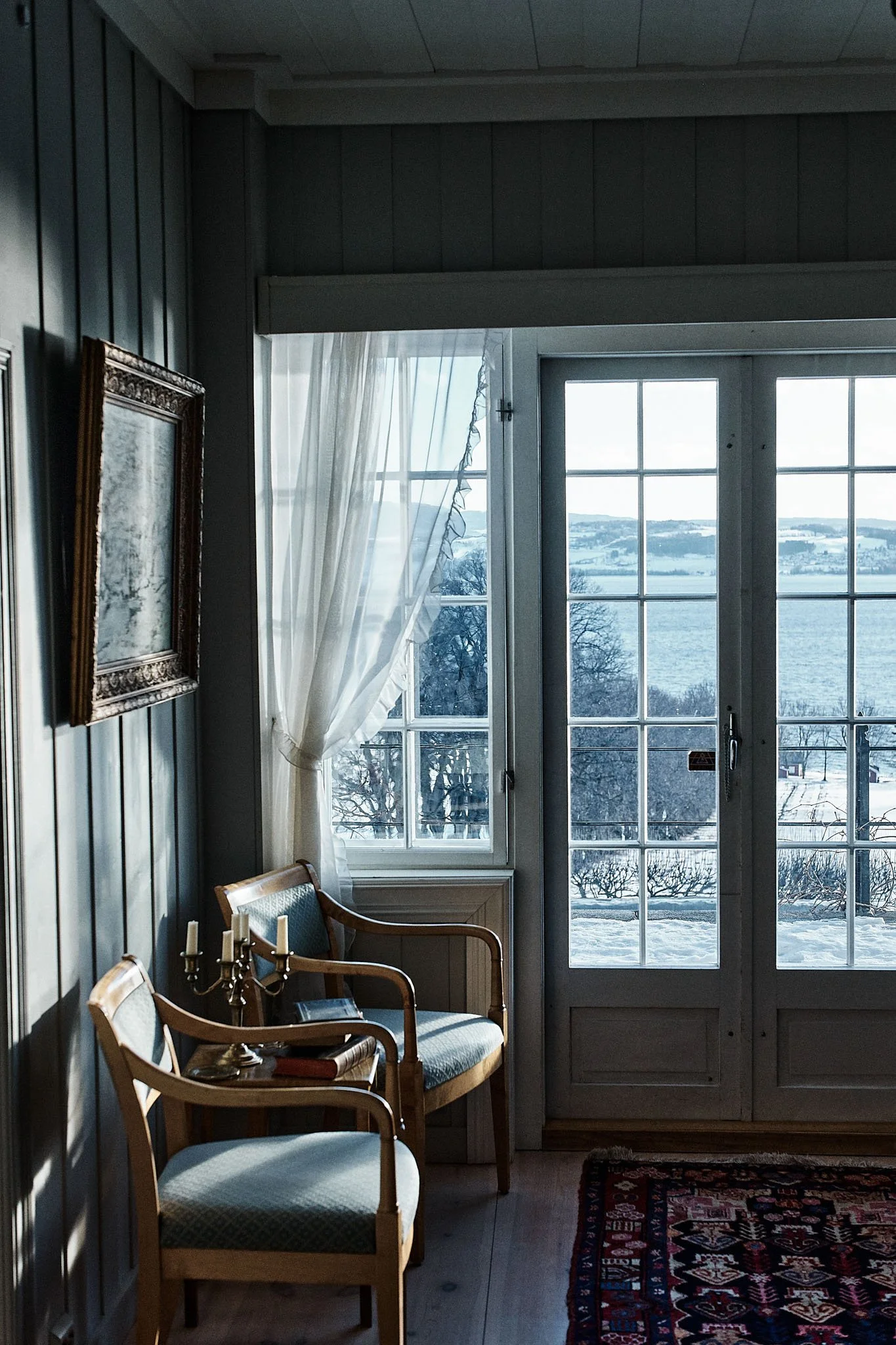 Sunlit interior room with two wooden chairs, a candelabra, and a window with white curtains overlooking a snowy landscape with trees and water.