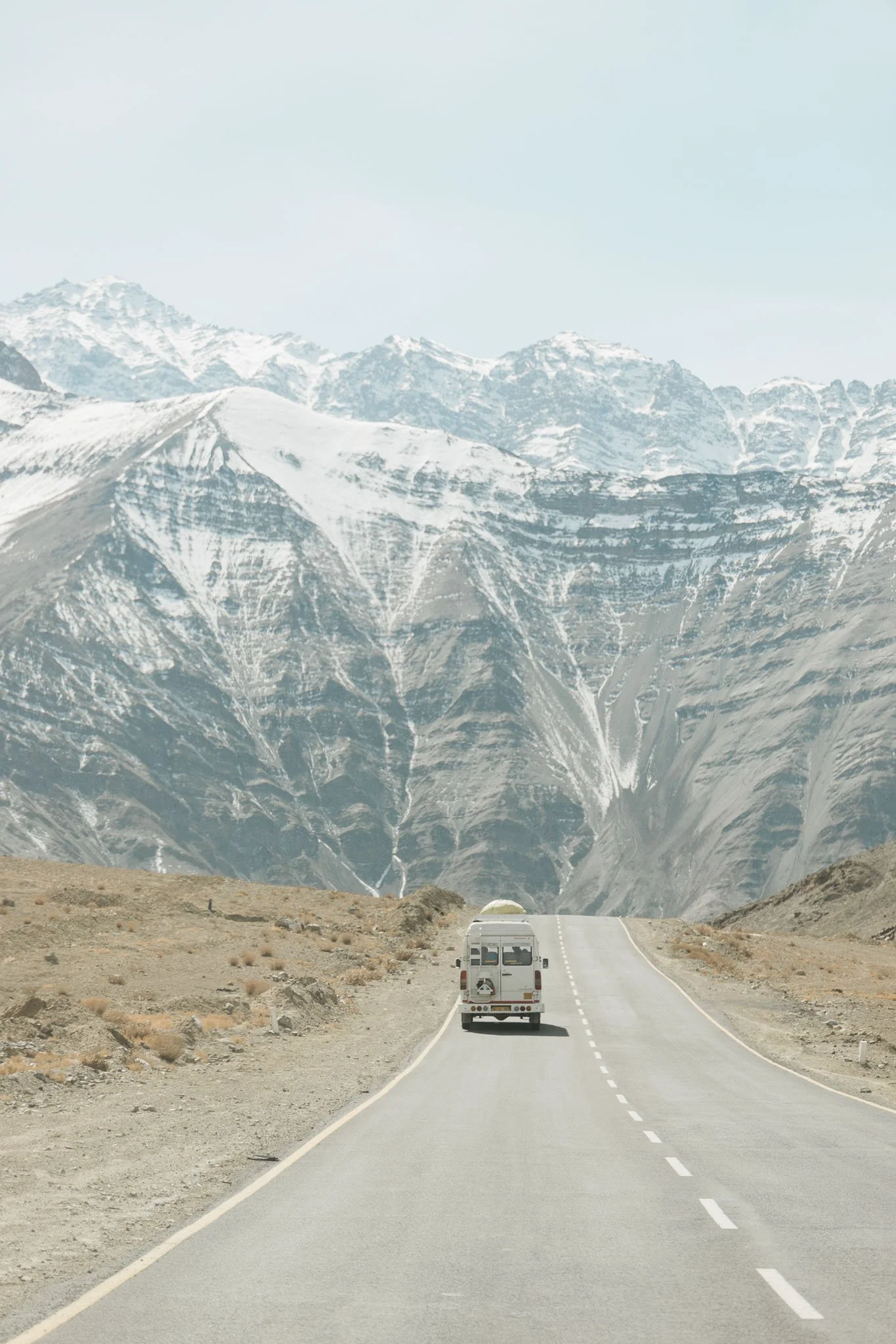 A white camper van driving on a straight mountain road through a barren desert landscape with snow-capped mountains in the background.