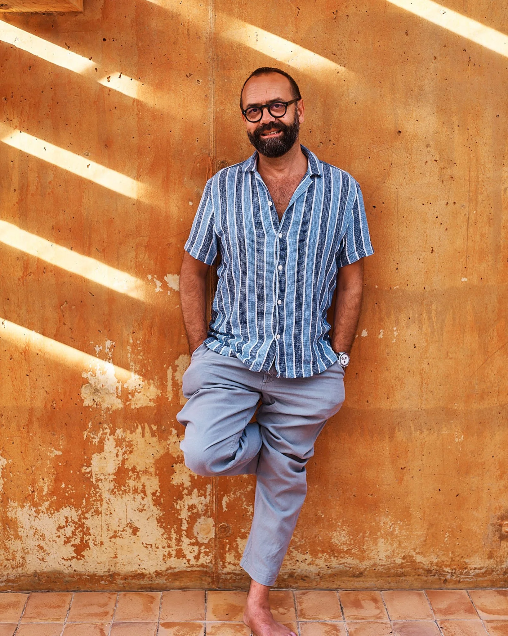 A man with a beard and glasses leaning against an orange wall, wearing a blue striped shirt and light gray pants with one foot raised and hands in pockets.