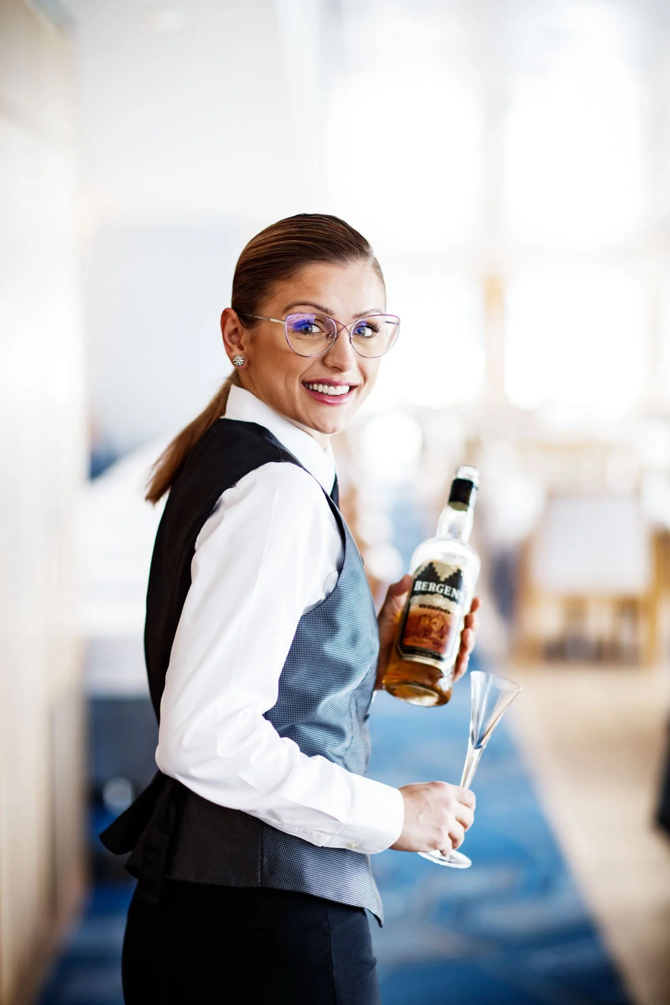 Smiling woman dressed as a bartender or server holding a bottle of Bergen's whiskey and a cocktail glass, standing in a bright indoor setting.