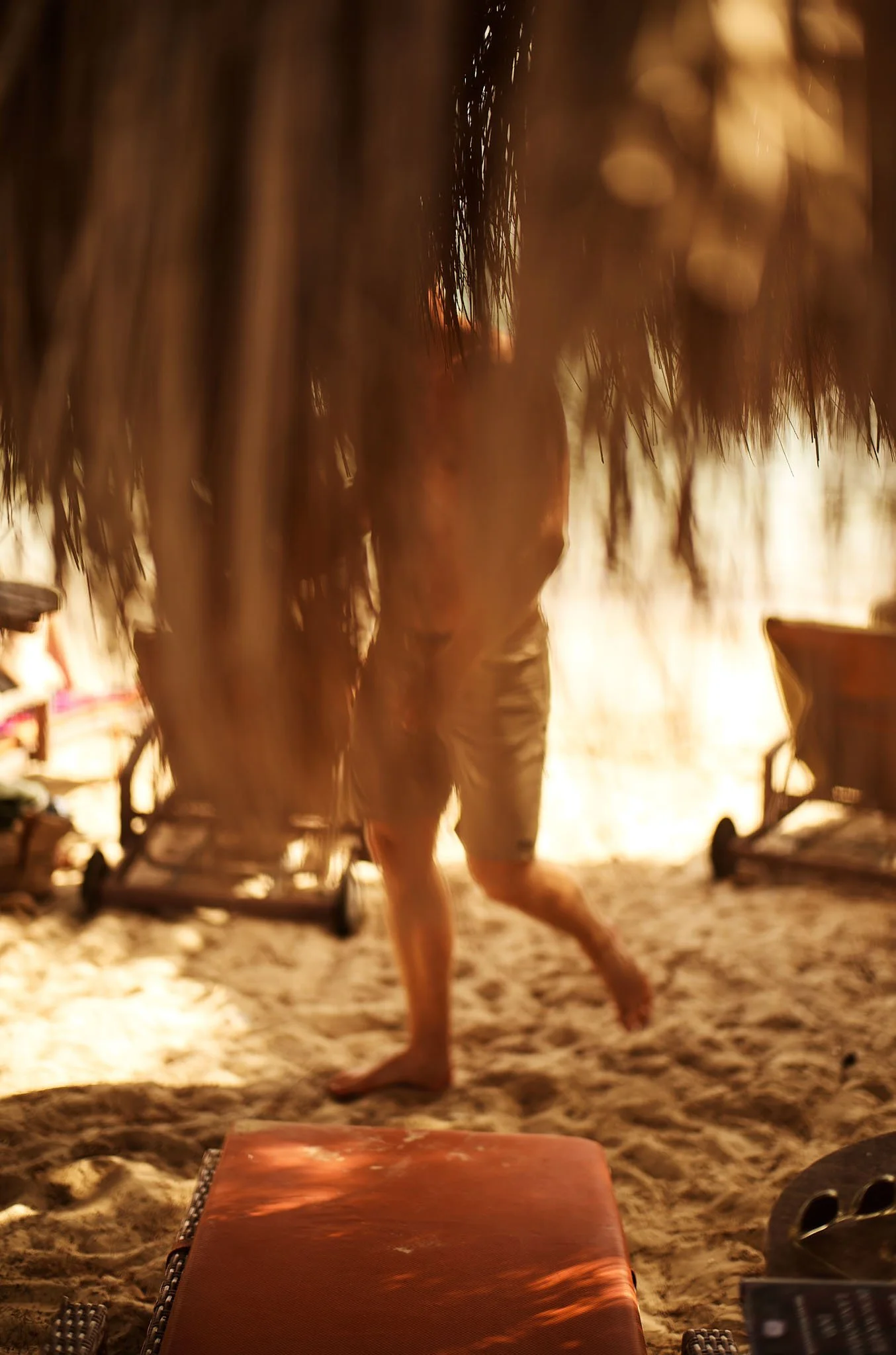 A person walking barefoot on a sandy beach, partially obscured by palm fronds from a thatched roof, with lounge chairs in the background.