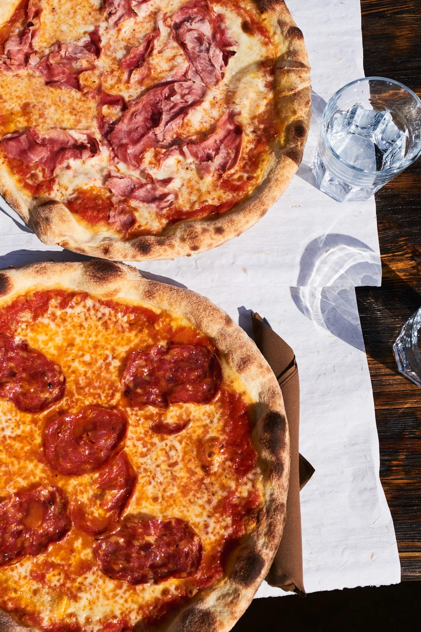 Two pizzas topped with cheese, with one also topped with ham and the other with pepperoni, on a white tablecloth with glasses of ice water.