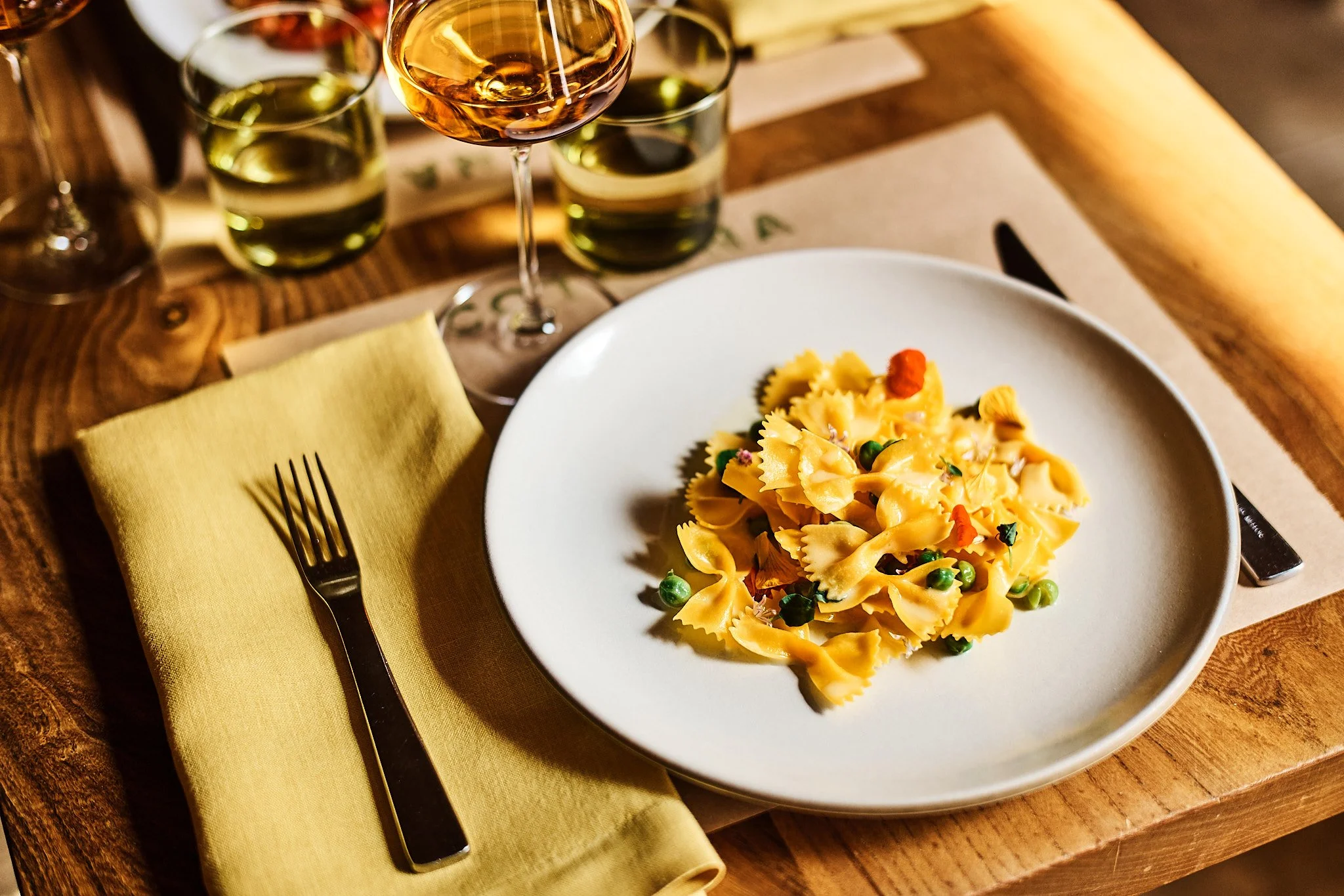 A dinner plate with bowtie pasta and green peas, garnished with herbs and a red pepper flake, on a wooden table with a yellow napkin and a fork. In the background are glasses of wine and water.