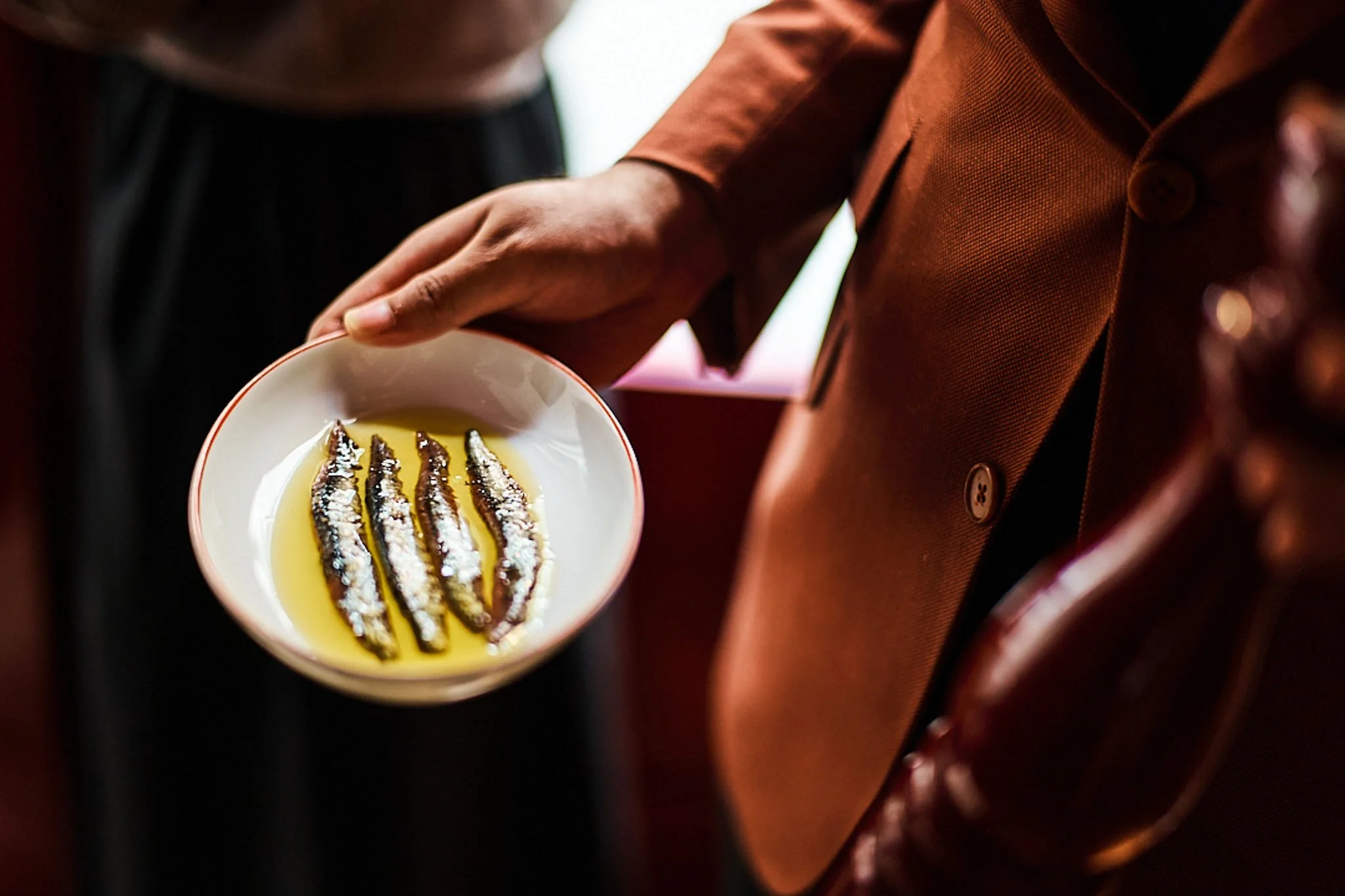 A person holding a white bowl with five small fish laid in olive oil, garnished with sea salt.