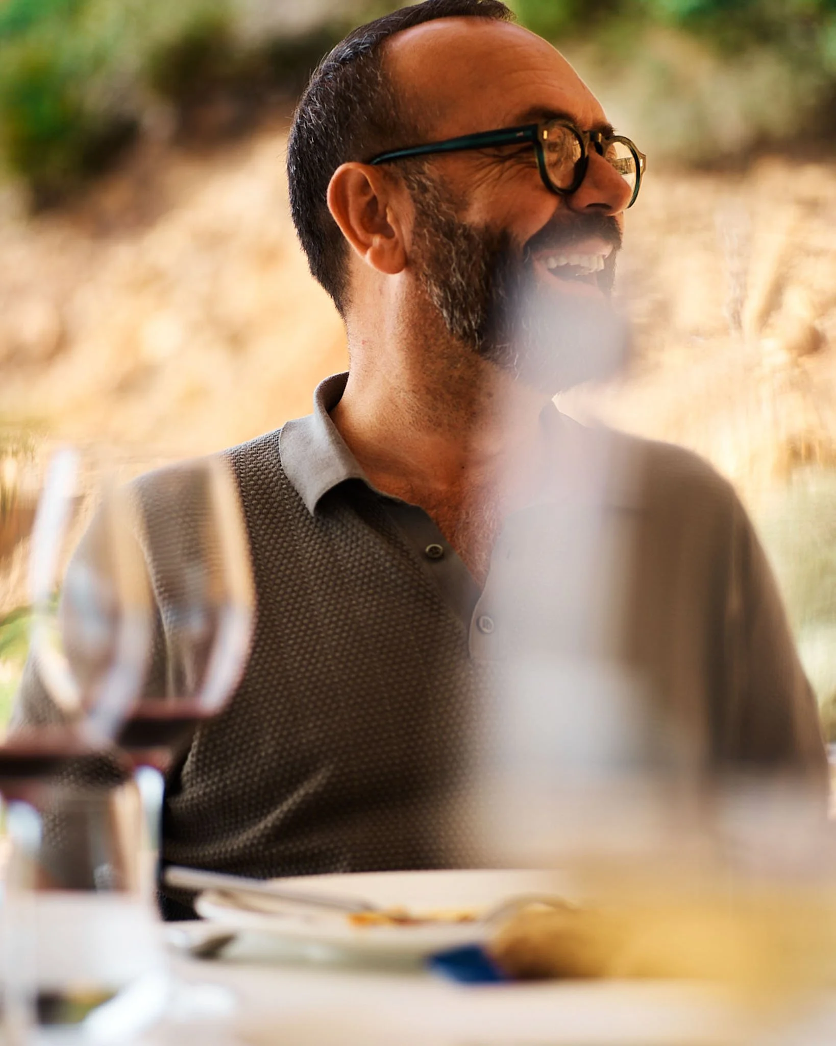 A man with short hair, glasses, and a beard, smiling and laughing while sitting at a table outdoors at a restaurant or cafe.