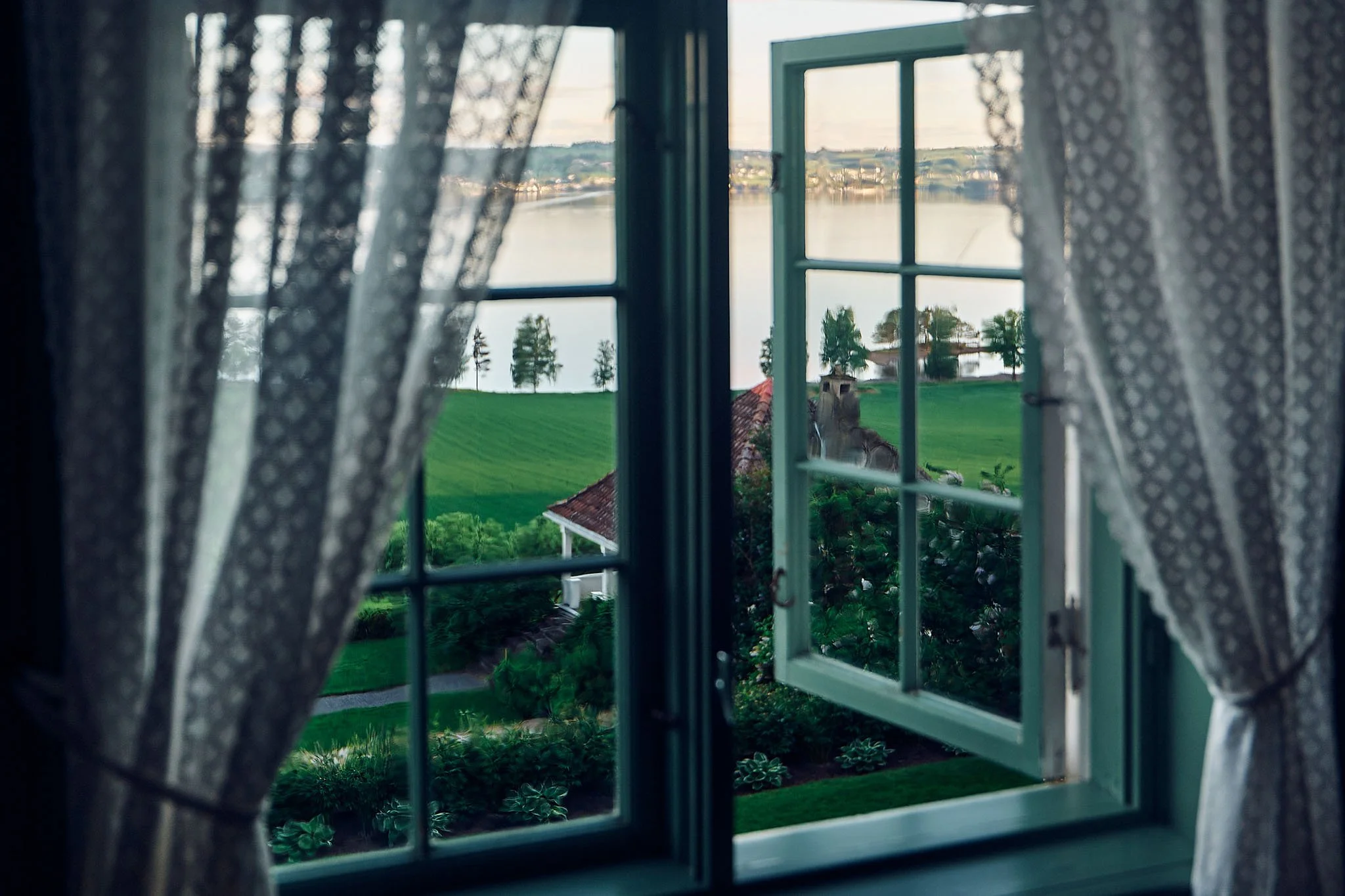 View through a window showing green grass, trees, a white building with a red roof, and a body of water with a distant shoreline.