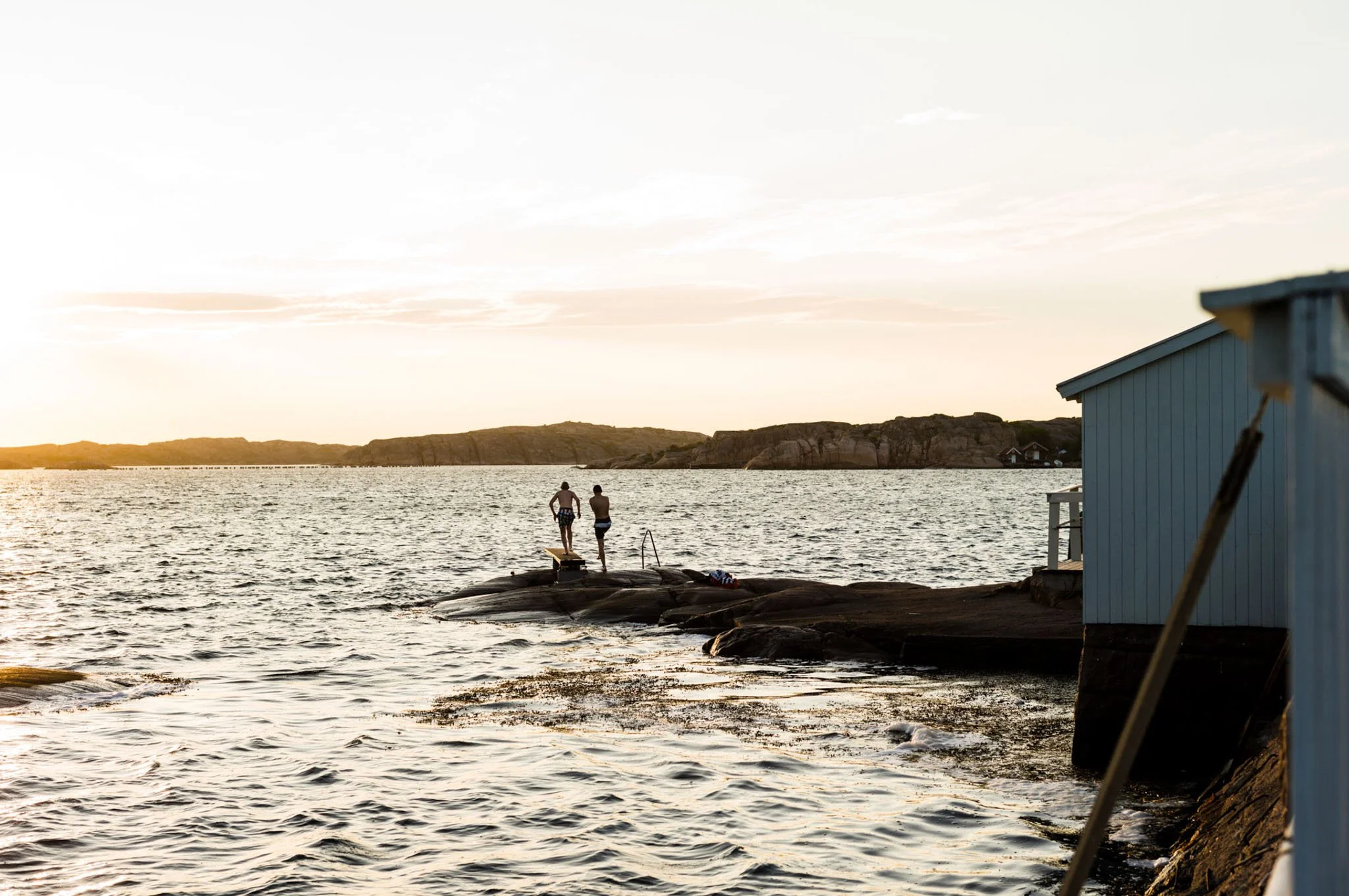 Two people standing on rocks by the water at sunset, with a small blue building on the right and distant hills in the background.