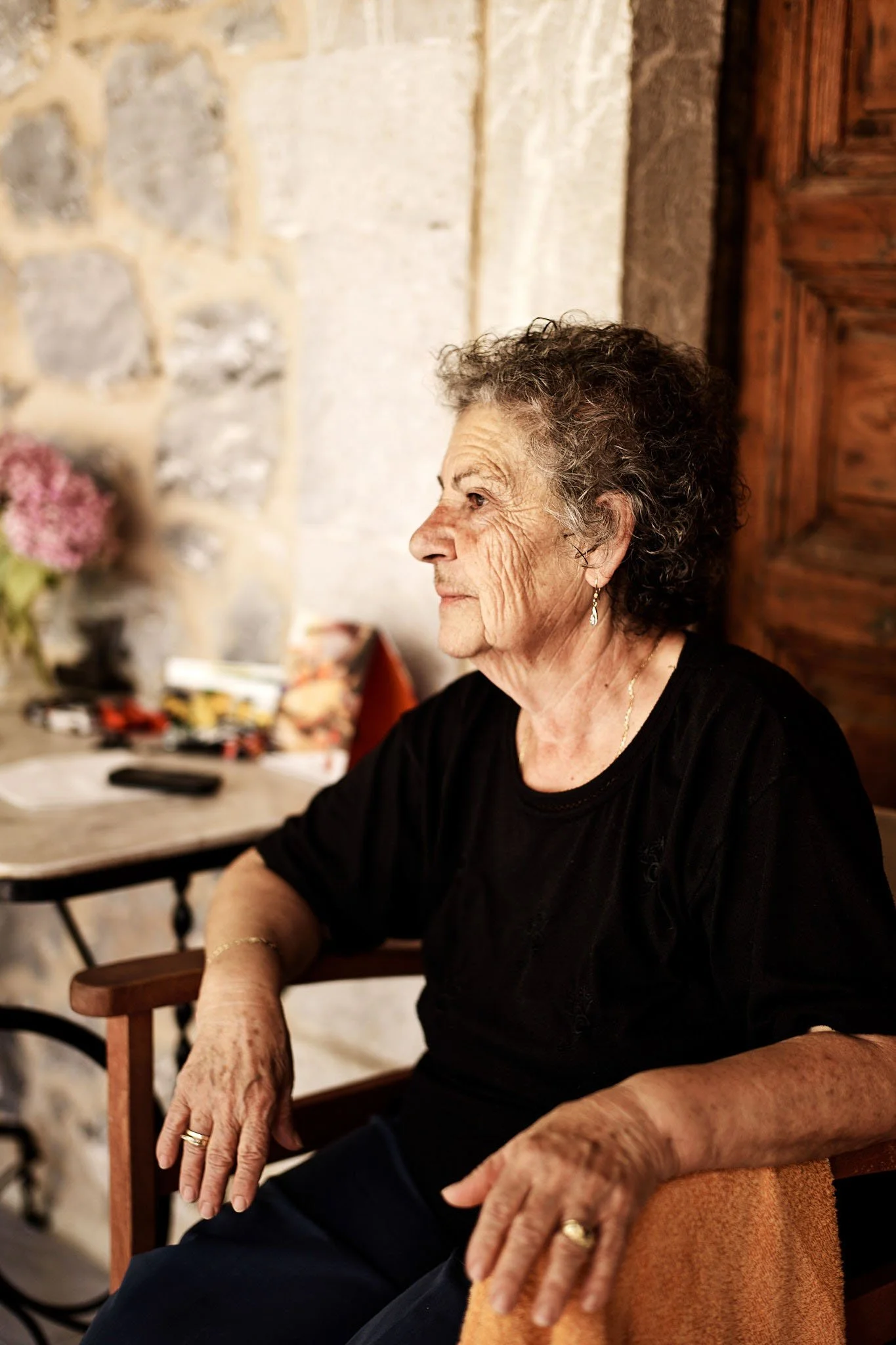 An elderly woman with curly gray hair wearing a black shirt and jewelry, sitting inside a room with stone walls, looking to the left.
