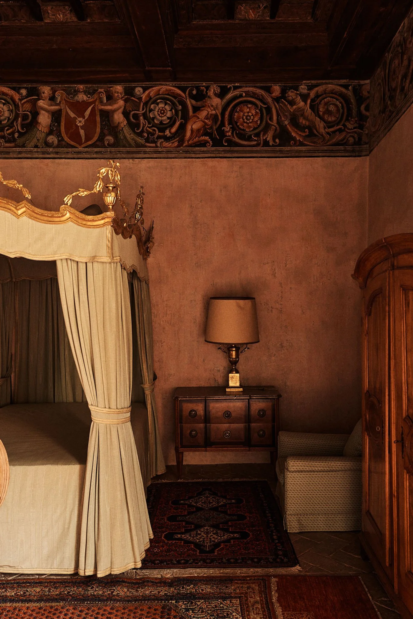 An elegant bedroom corner with a four-poster bed with cream-colored curtains, a vintage wooden cabinet with a table lamp on top, a cozy armchair, and decorative wall molding featuring cherubs and floral motifs.