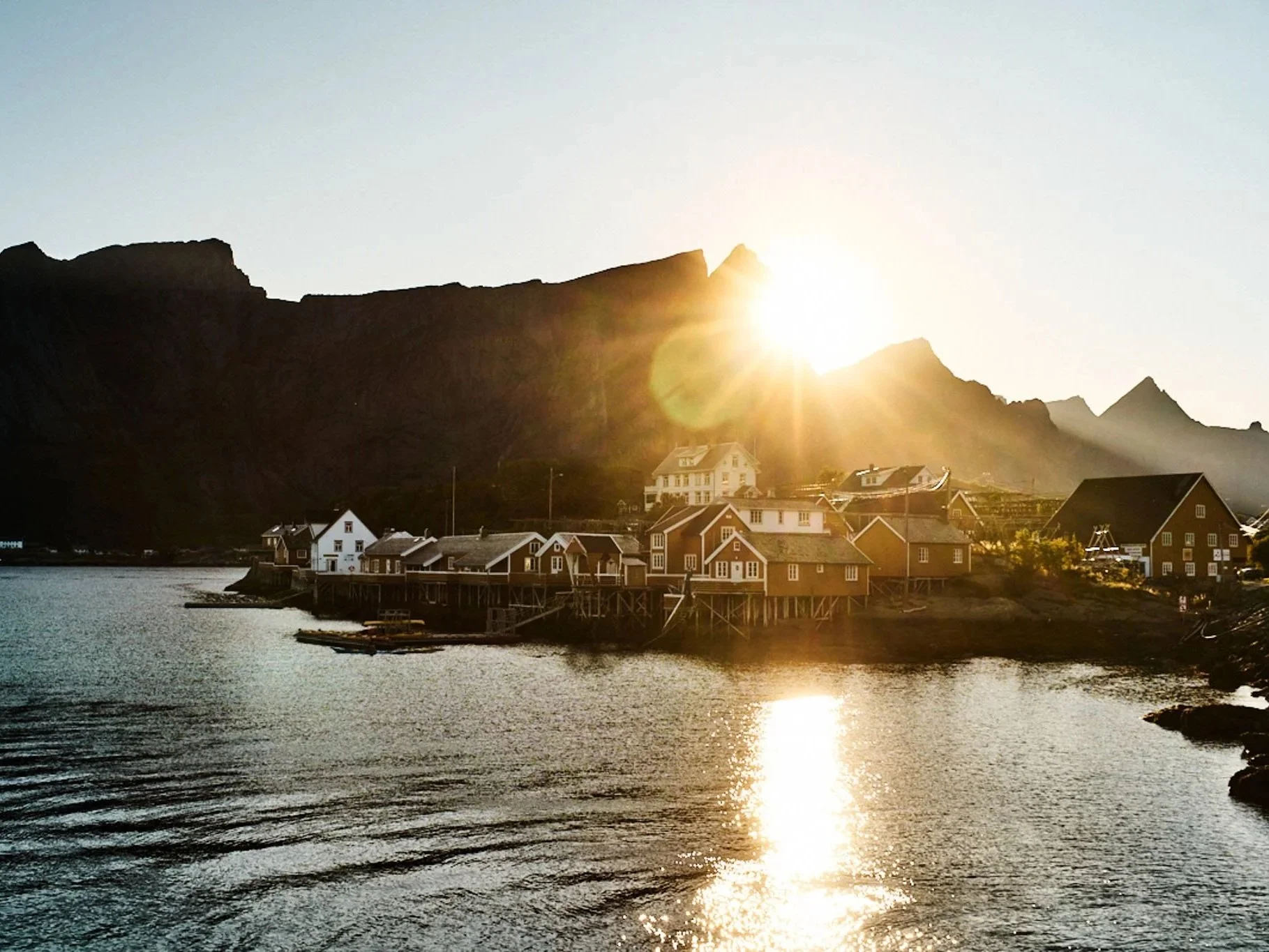 Sunset over a small village with houses on stilts beside a body of water and mountains in the background.