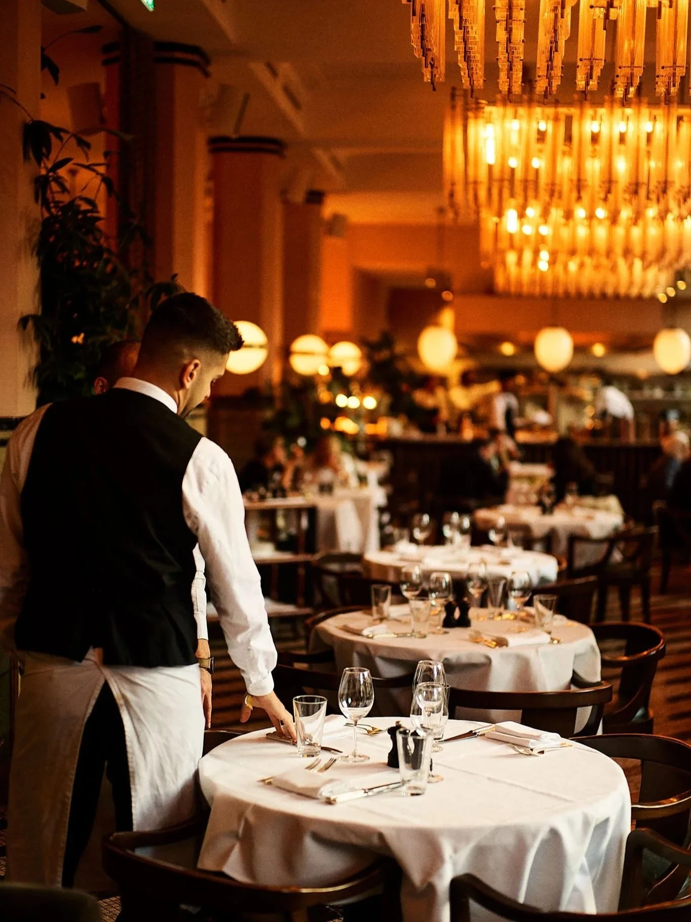A waiter setting a table in a dimly lit upscale restaurant with round tables, white tablecloths, glasses, and utensils. The restaurant has warm lighting and chandeliers.