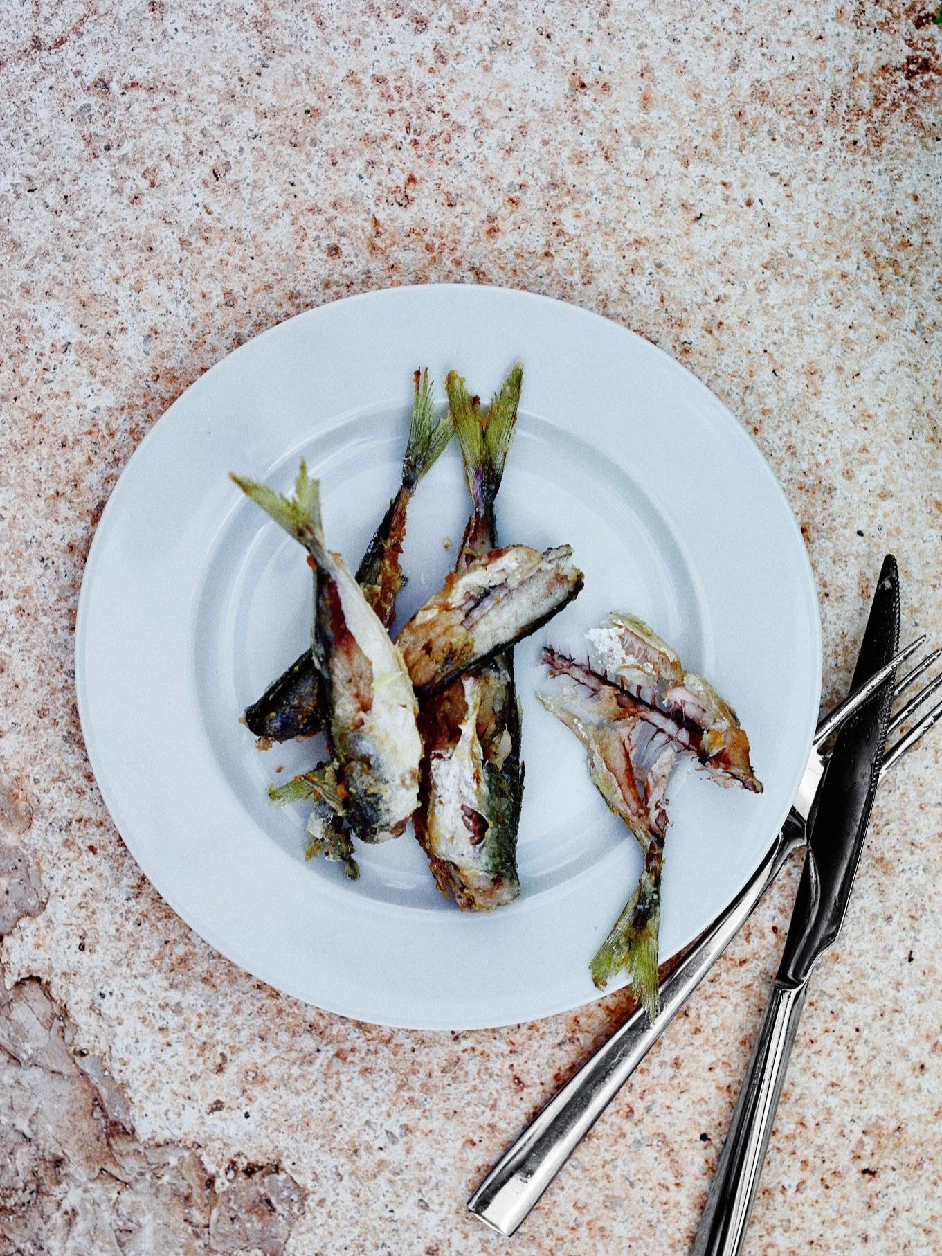 A white plate of small grilled fish with charred tails, placed on a granite countertop with a fork and knife nearby.