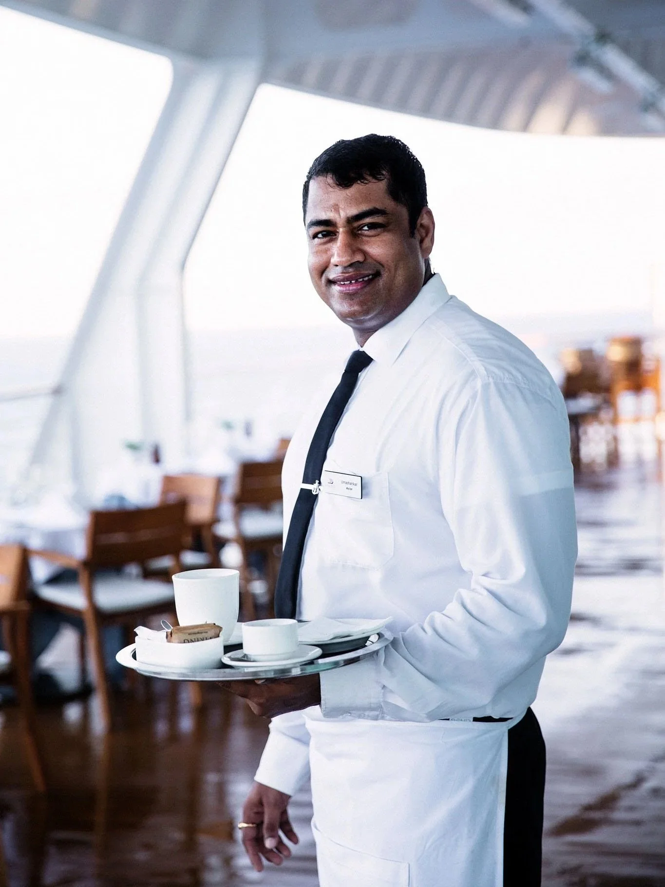 A smiling male waiter in a white shirt, black tie, and apron holding a tray with coffee cups in a restaurant or hotel with open-air, modern design and ocean view.
