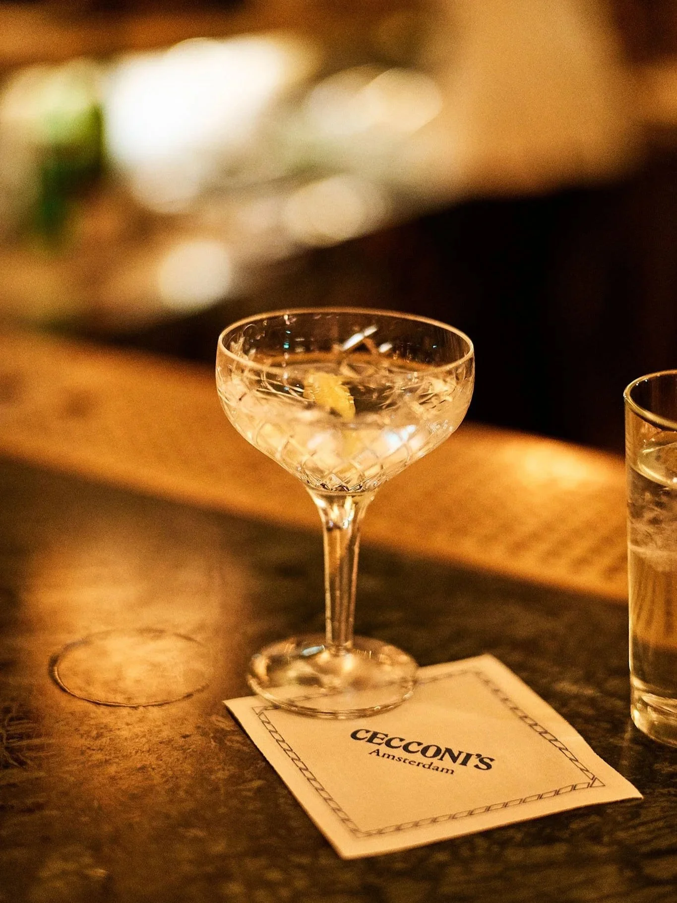 A cocktail glass on a bar counter with a lemon twist garnish, placed on a napkin with 'Ceccotti's Amsterdam' written on it, next to a shot glass of water.