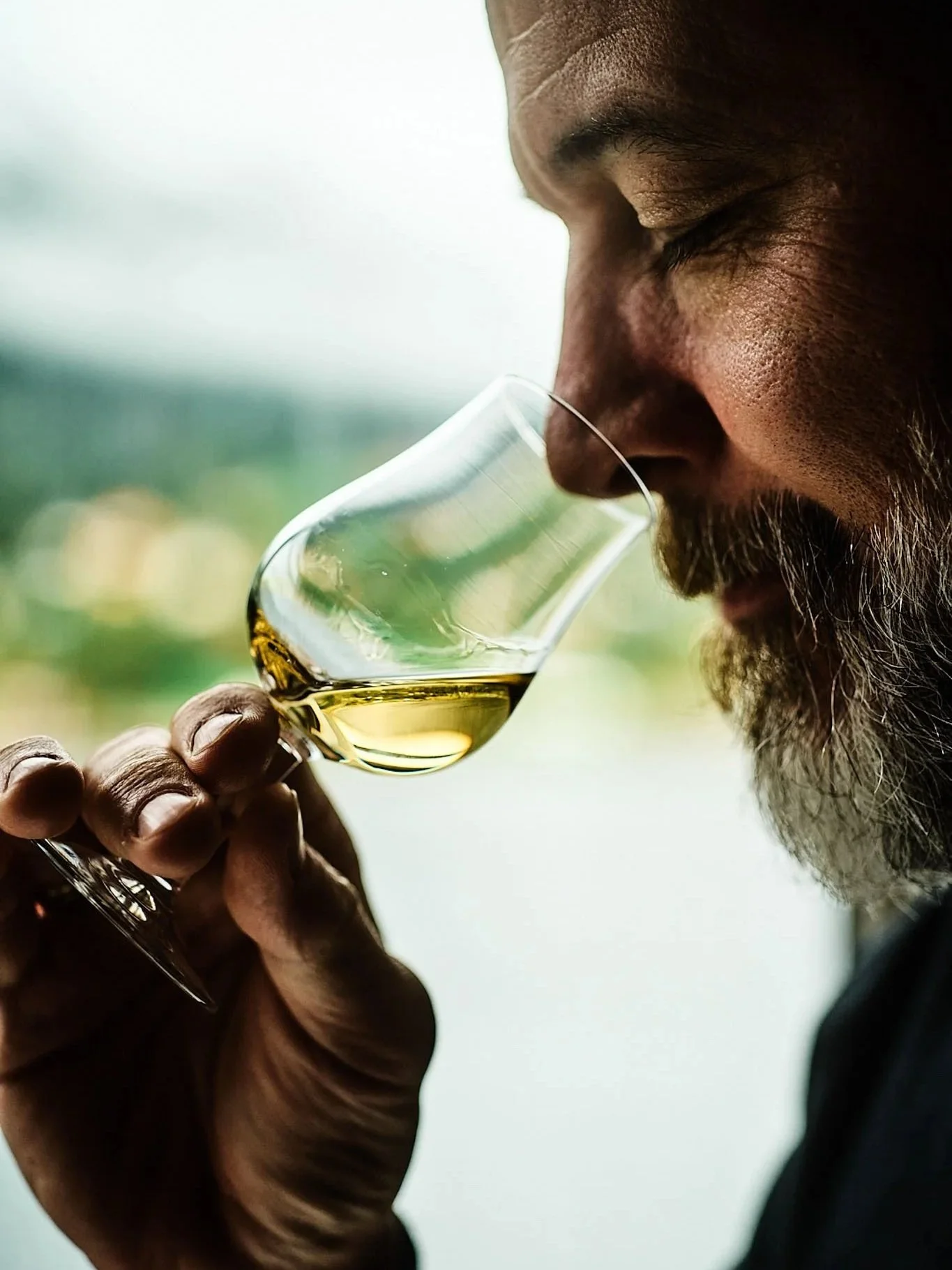 A man with a beard and closed eyes holding a glass of yellowish liquor close to his nose, appearing to smell the drink.