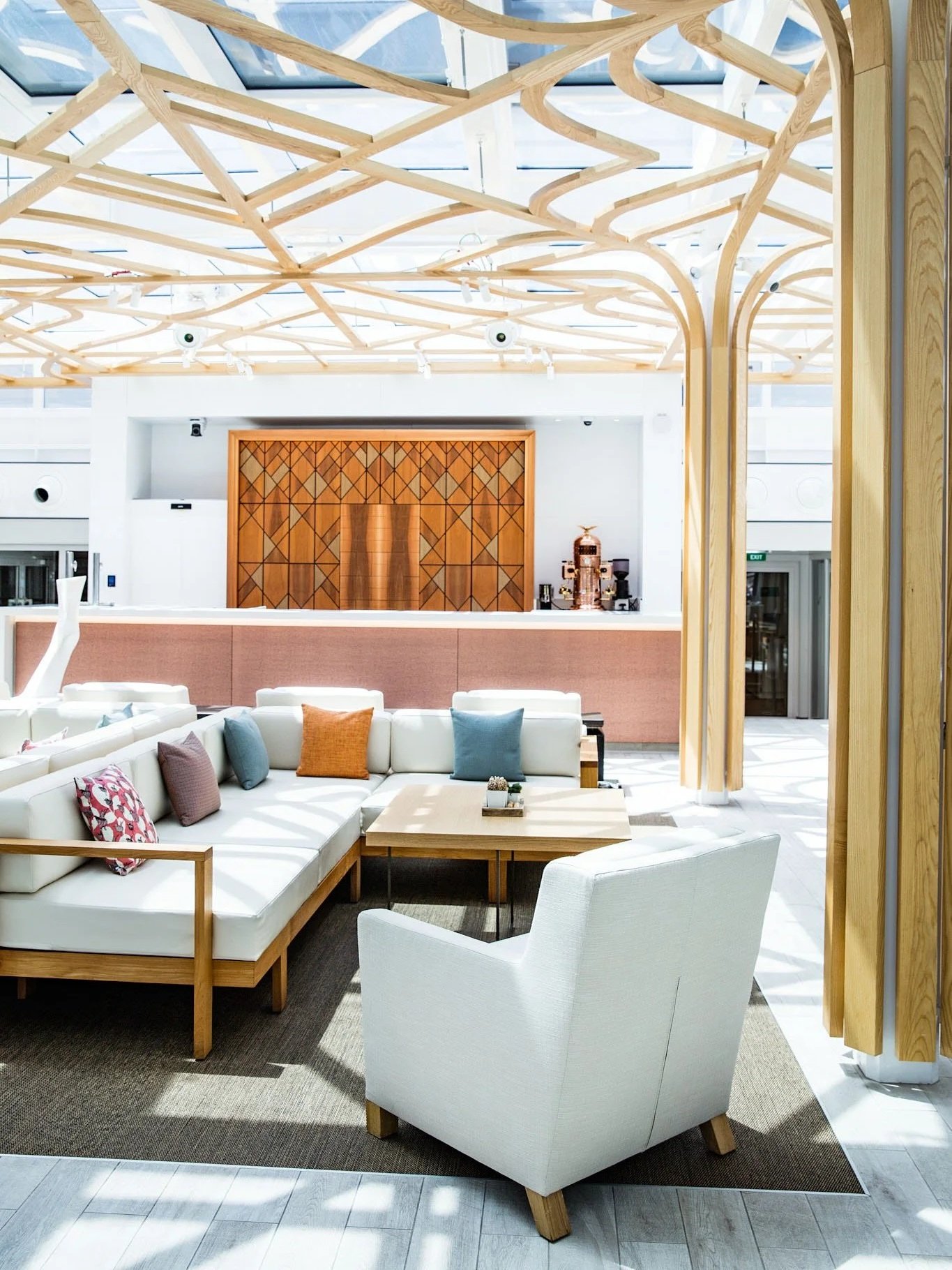 Modern hotel lobby with white seating, colorful pillows, wooden accents, and intricate wooden ceiling design.