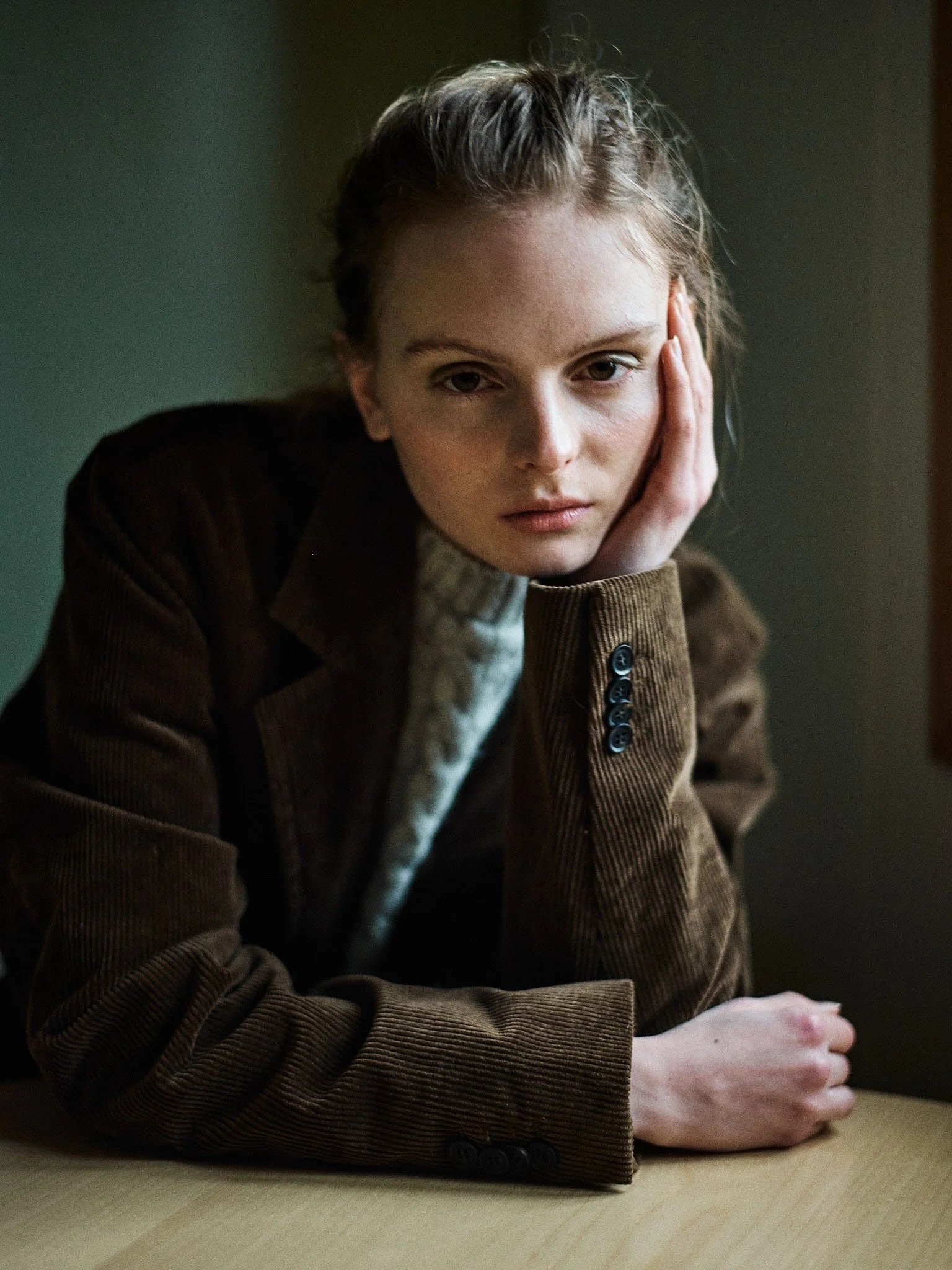 Young woman with blonde hair sitting at a table resting her head on her hand, wearing a brown corduroy blazer and a patterned turtleneck in a dimly lit room.